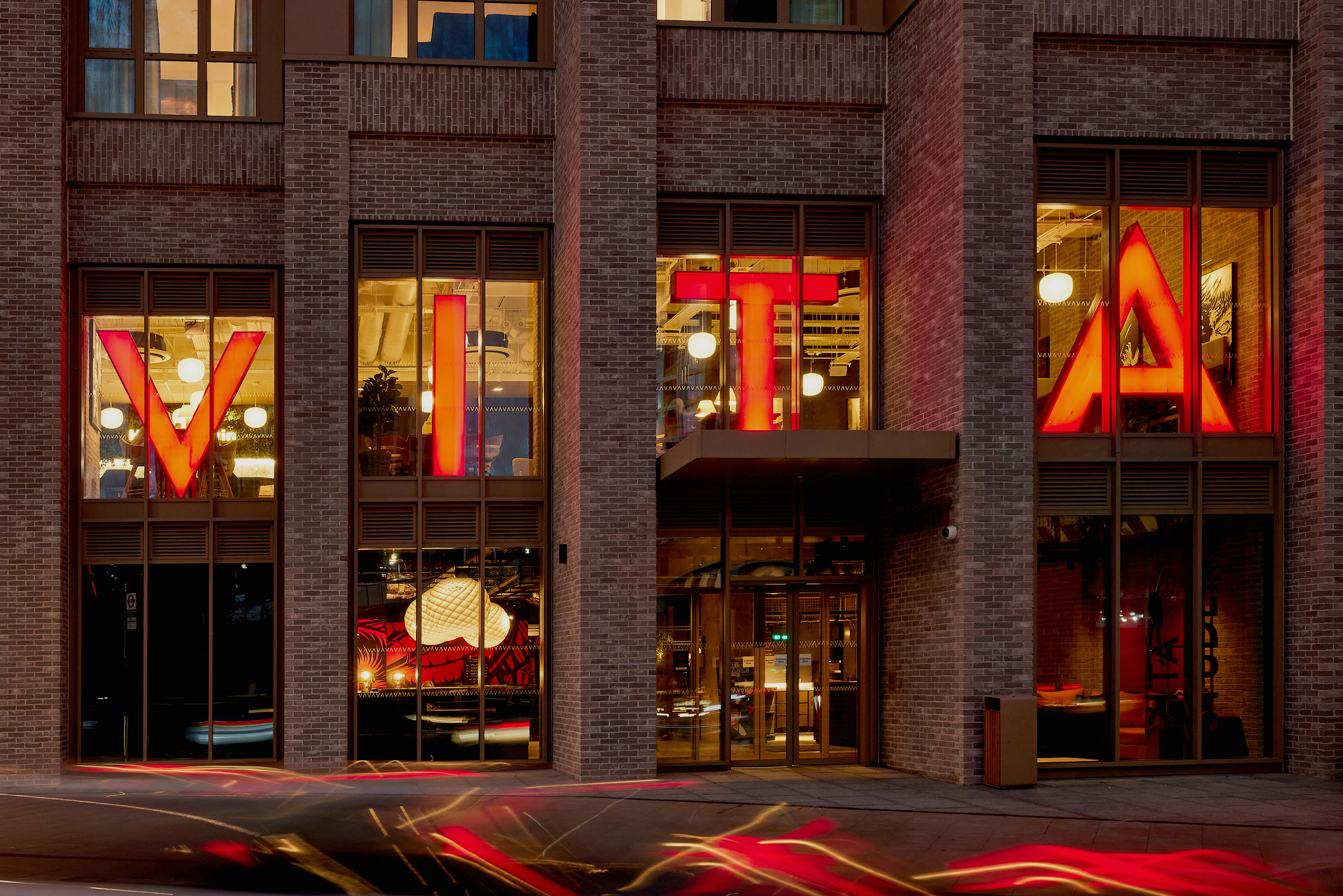 The brick building with large vertical windows displays the illuminated red letters "VITA." Inside, modern decor abounds with bright lights and bold artwork. In the foreground, blurred streaks of red and white suggest UK traffic bustling through the vibrant scene.