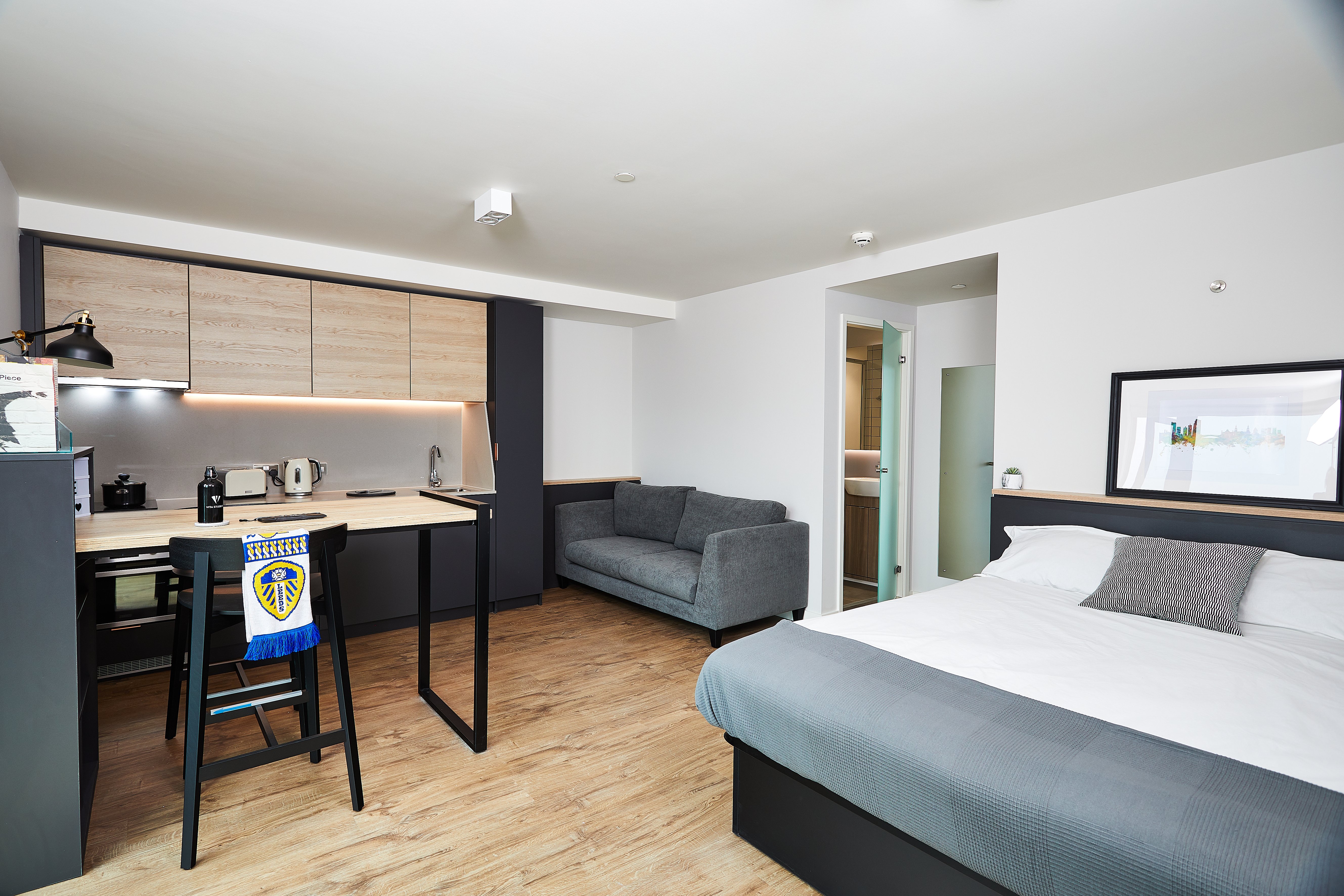 Modern studio apartment with a wood floor, featuring a bed with gray bedding, a gray sofa, and a compact kitchenette. A high chair with a sports towel, a microwave, and a kettle are on the counter. A bathroom is visible through an open door.