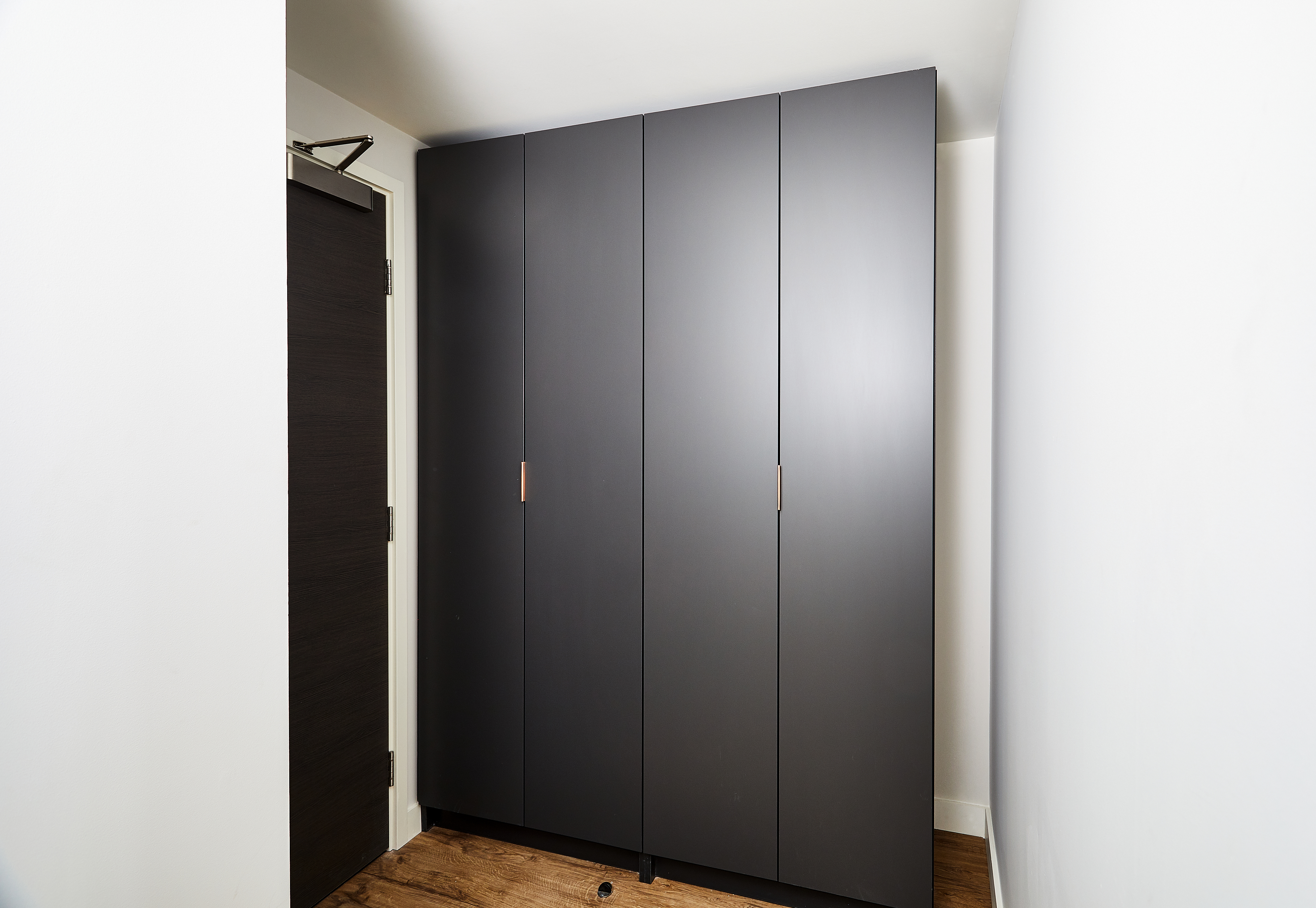 A narrow room with a matte black wardrobe against the wall. The wardrobe has sleek, flat panels and minimalistic handles. A dark wooden door is partially visible on the left, set against white walls and a wooden floor.
