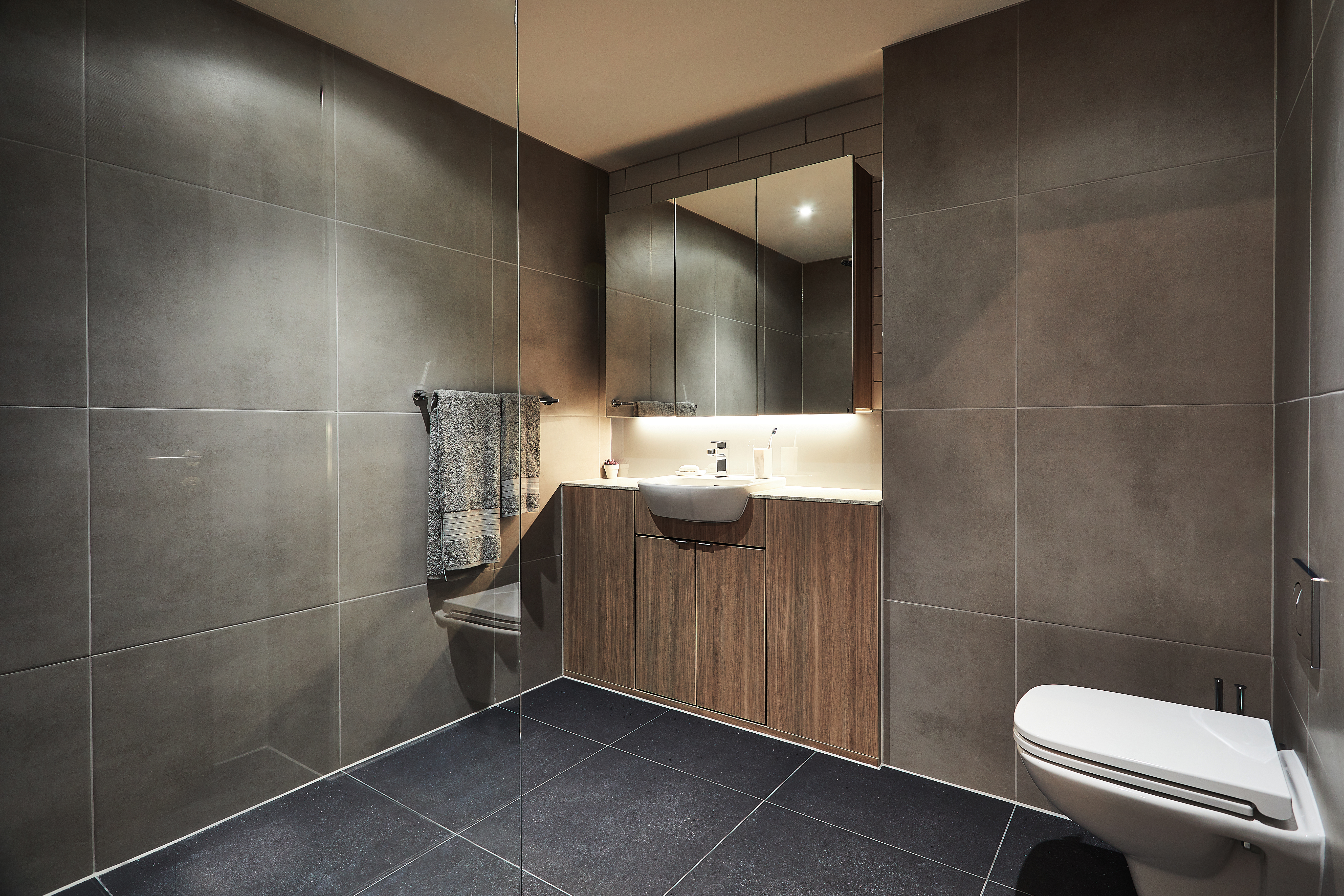 Modern bathroom with dark tiled walls and floor. Features a wall-mounted sink with a wooden vanity, a mirror, a towel hanging on a rail, and a toilet. Soft lighting under the mirror creates a warm ambiance.