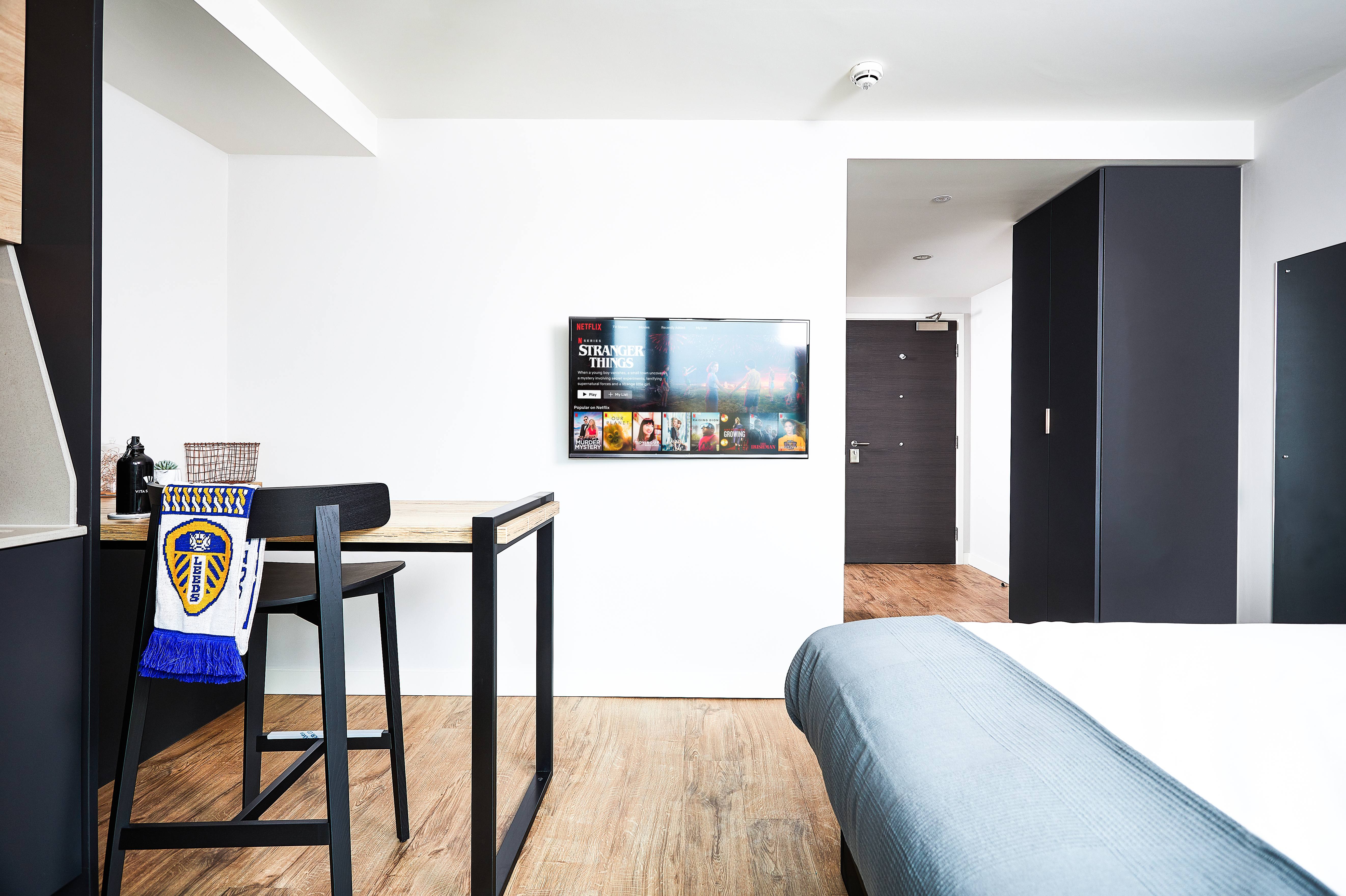 Modern hotel room with wood flooring, a bed with a blue blanket, a wall-mounted TV displaying a streaming service, and a wooden table with two high stools. A sports team scarf hangs from a stool. The entrance door is visible in the background.