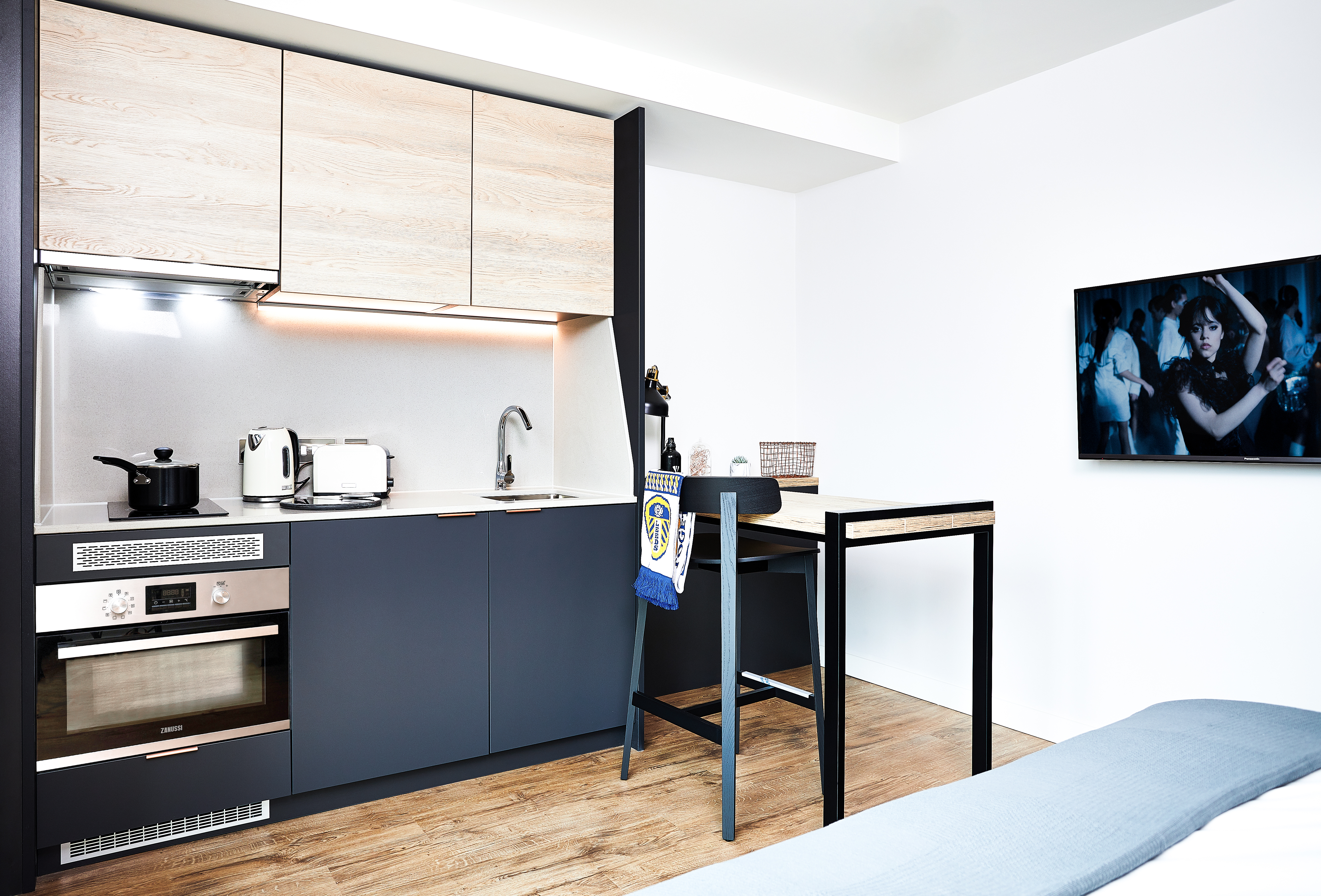Modern kitchenette with wood and black cabinetry, silver appliances, and a small bar-style table. A pot, toaster, and kettle are on the countertop. A wall-mounted TV displays an action scene. Light wood flooring and a section of a gray bed are visible.