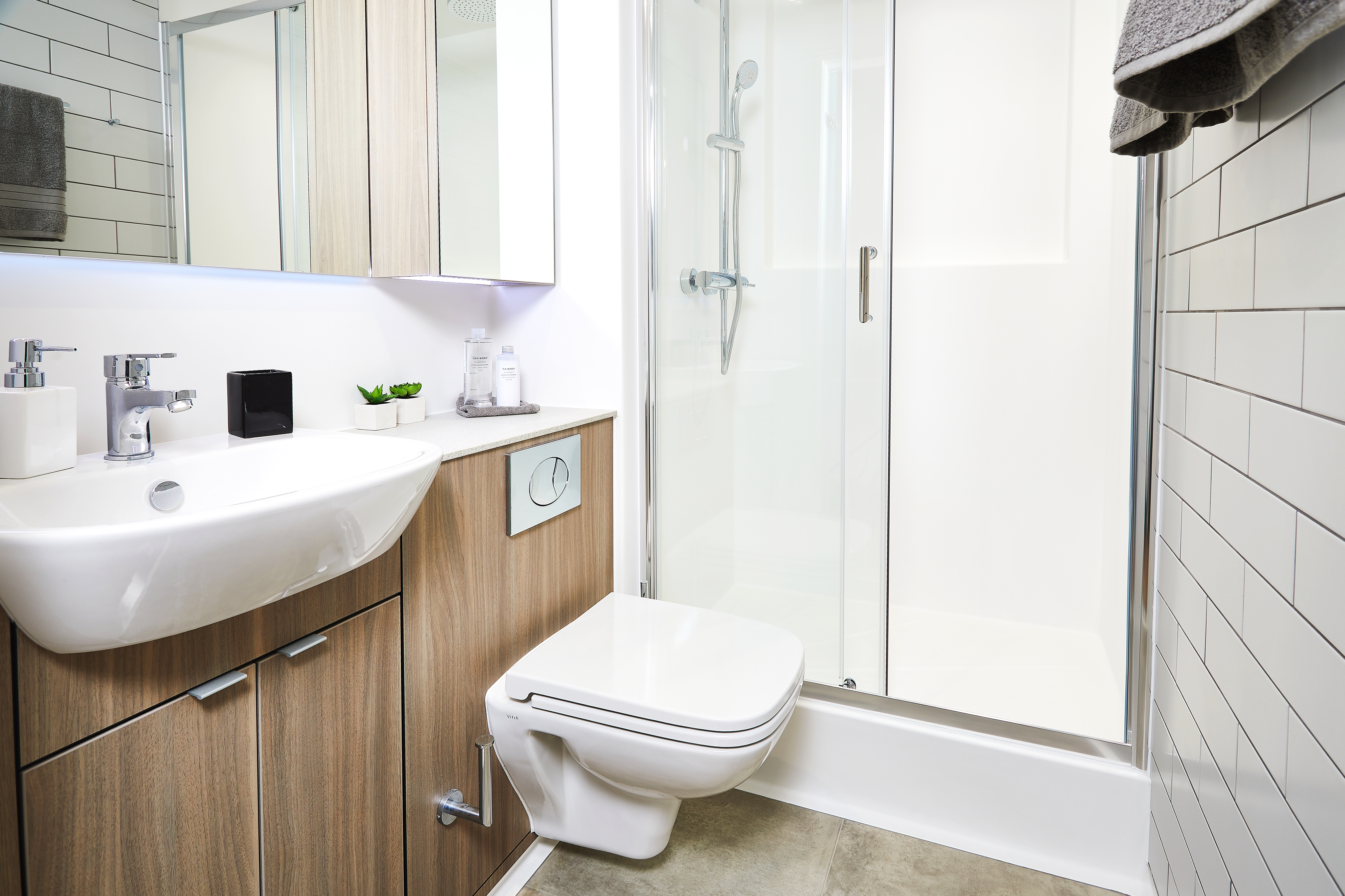The modern bathroom on Station Street features a white sink, wooden cabinetry, and a wall-mounted toilet. A glass-enclosed shower with a chrome showerhead enhances the elegance. White tiled walls add brightness, while a small plant and soap dispensers sit neatly on the counter.
