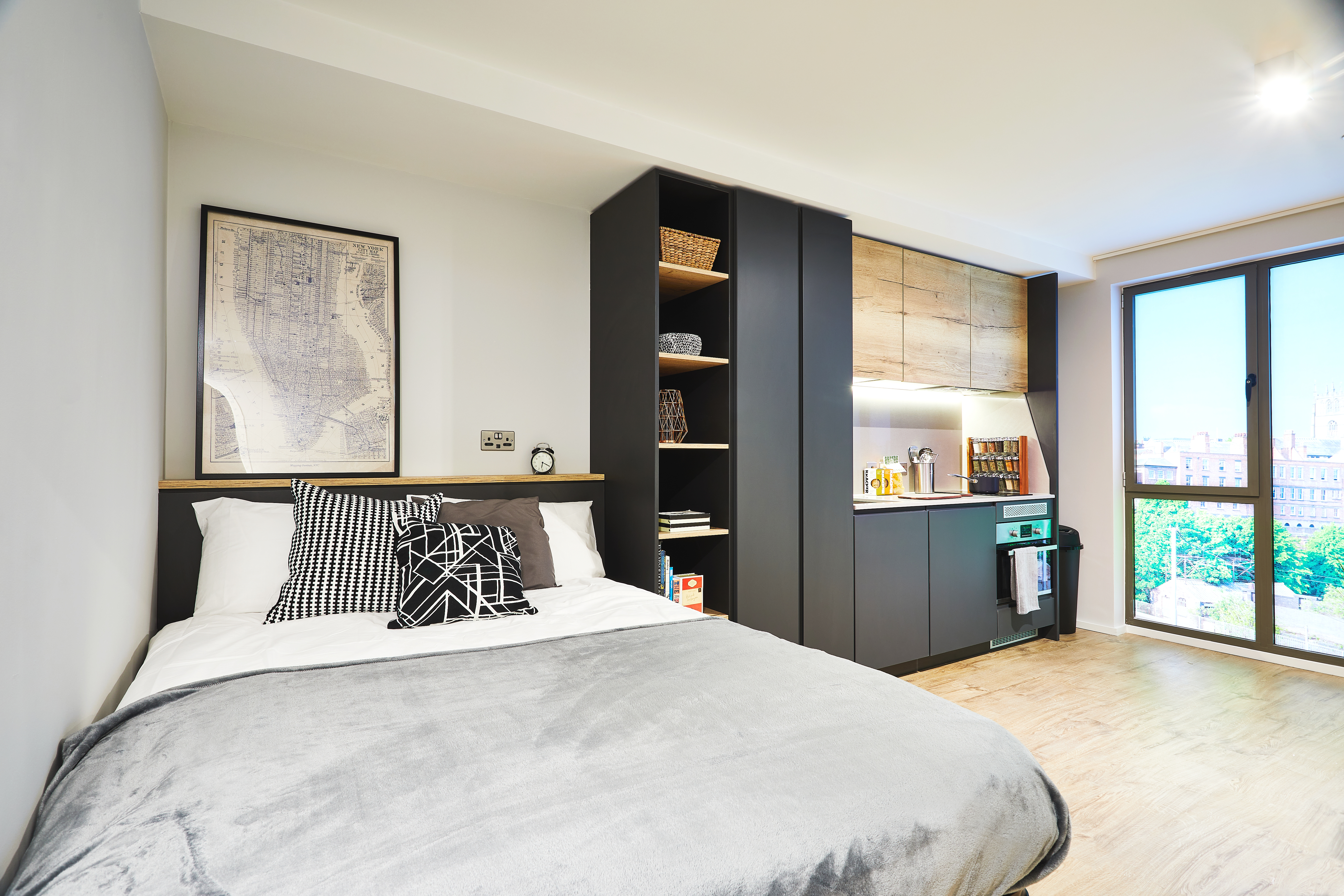 A modern bedroom on Station Street features a neatly made bed with black and white pillows and a gray blanket. A map is framed on the wall, opposite dark cabinets and decor-filled shelves. A large window reveals a breathtaking view of the cityscape and blue sky.