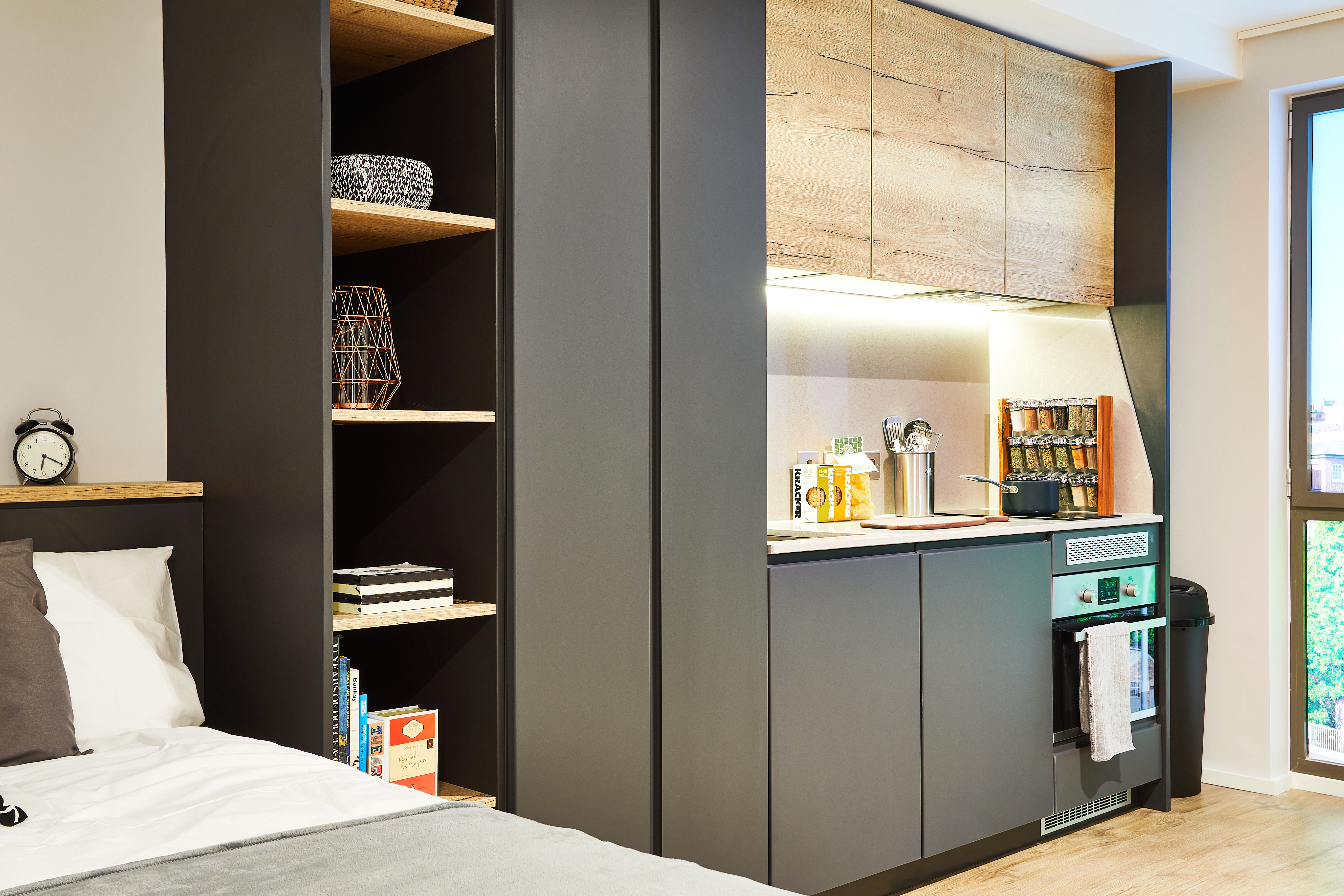 Nestled on Station Street, this modern studio apartment boasts a sleek kitchenette with black cabinets, wood accents, and stainless steel appliances. Open shelving displays books and decor items. A neatly made bed peeks from the left, while a window bathes the space in natural light.