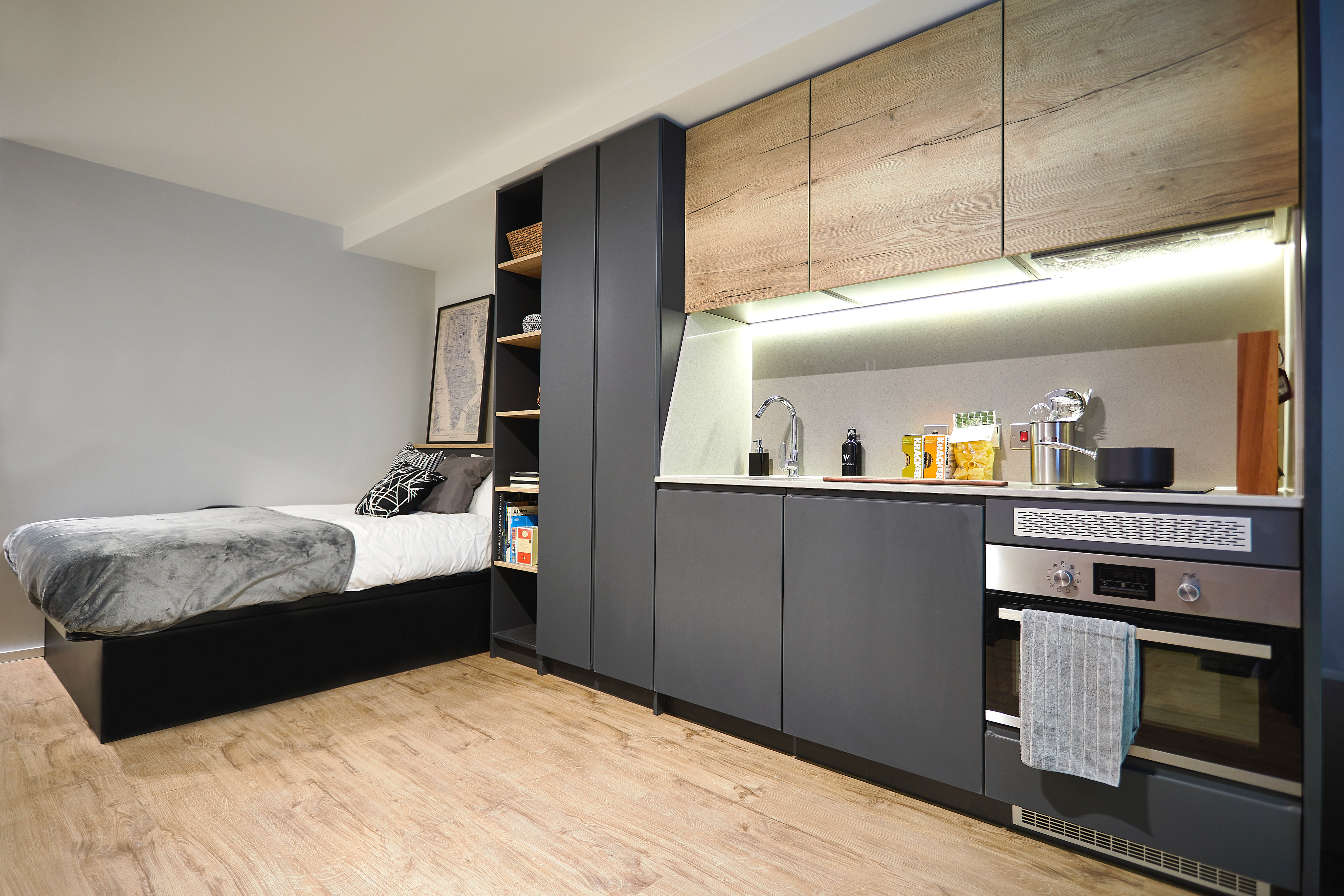 Located on Station Street, this modern studio apartment boasts a wooden floor and features a compact kitchen with dark cabinets, an oven, and various kitchen essentials. The bed, adorned with gray bedding, sits adjacent to the kitchen, complemented by shelves filled with books and decor.