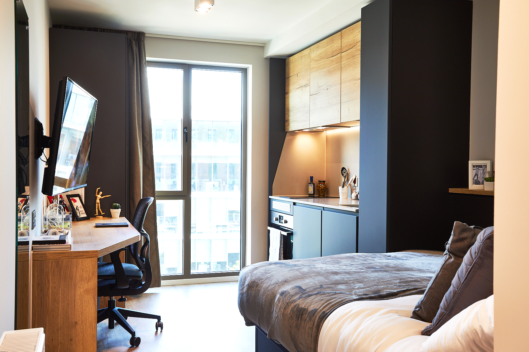 Located on Station Street, this modern studio apartment features a cozy bed on the right and a desk with a chair and TV on the left. Dark accents complement wood paneling, while a large window bathes the space in natural light. The compact kitchen area includes shelves adorned with decorative items.