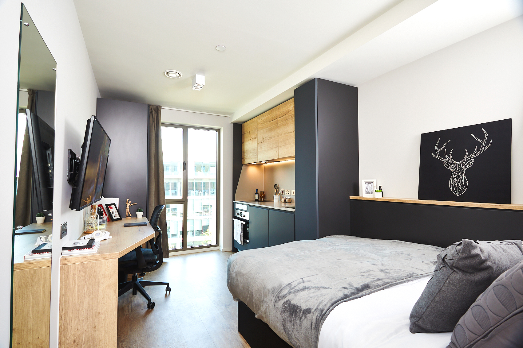 Nestled on Station Street, this modern bedroom features a cozy bed with a gray blanket, a wooden desk with a computer, and a wall-mounted TV. A black cabinet with wooden shelves stands proudly on the left, while stag artwork adorns the wall. Large windows with curtains illuminate the room.