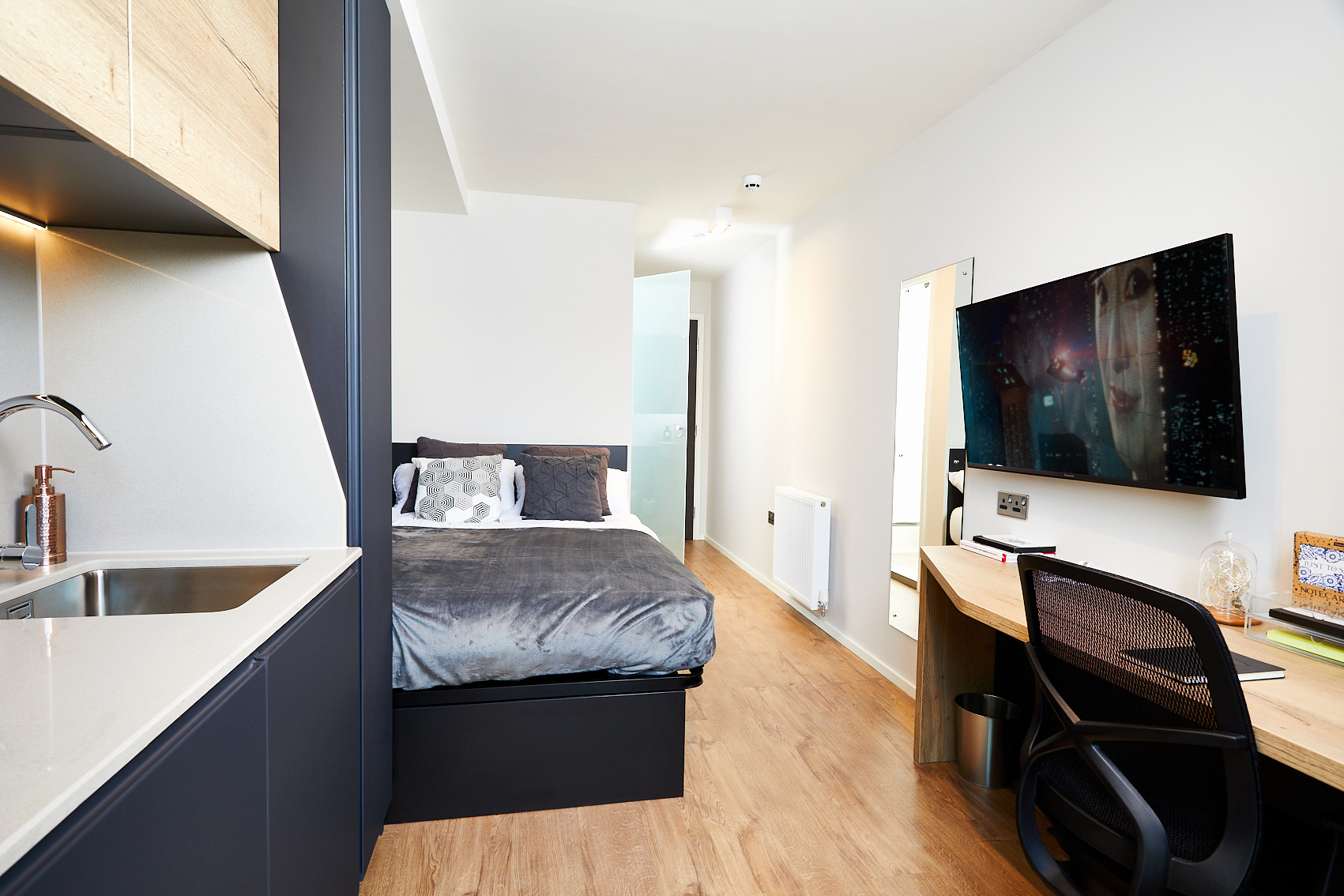 Nestled on Station Street, this modern studio apartment features wooden flooring, a cozy bed with gray bedding, a wall-mounted TV, and a sleek desk with black chair. A small kitchenette graces the left side while a long hallway leads to the bathroom in the background.