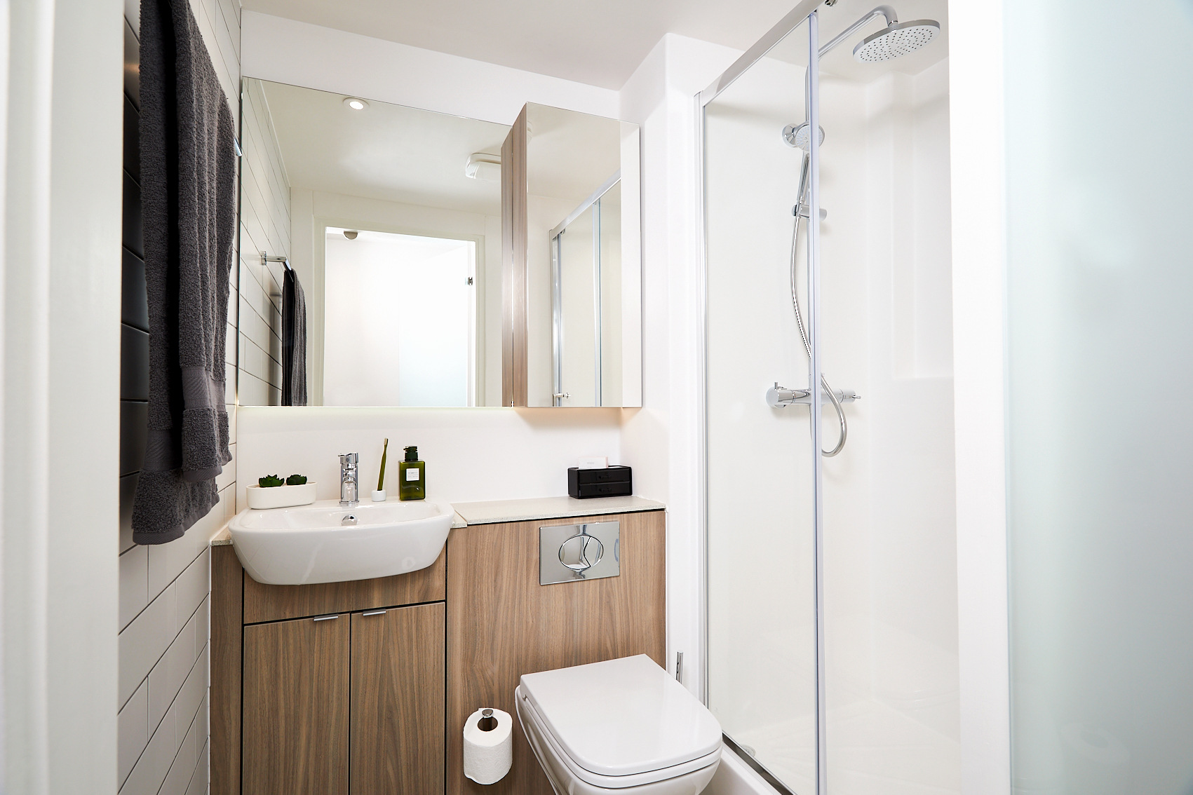 Modern bathroom on Station Street featuring a glass-enclosed shower, wall-mounted sink, and toilet with wooden cabinetry. A large mirror above the sink reflects light, enhancing the minimalist decor. White walls and a neatly folded towel complete the clean, sleek look.