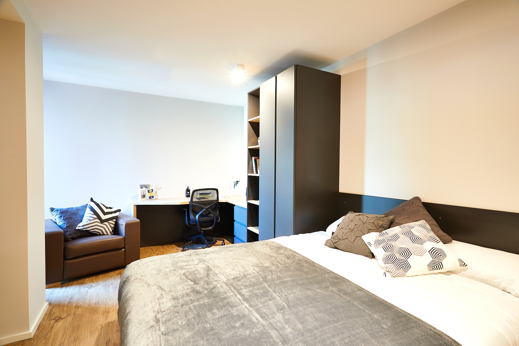 A bright, modern bedroom on Station Street features a double bed adorned with gray and patterned pillows and a soft gray blanket. A black shelving unit, a small desk with a swivel chair, and a cozy armchair by the window enhance the room's charm, while the hardwood floor adds warmth to the space.