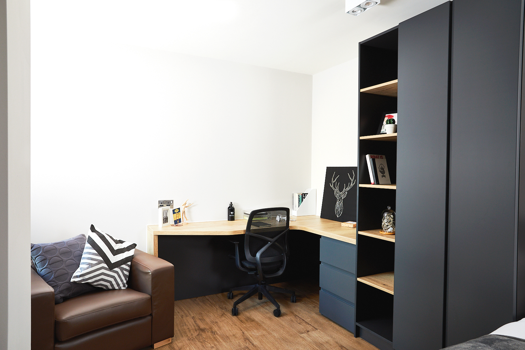 A modern home office on Station Street features a wooden L-shaped desk, a black office chair, and shelves displaying books and decor like a stag print. A brown leather armchair with patterned cushions stands against white walls and wood flooring.