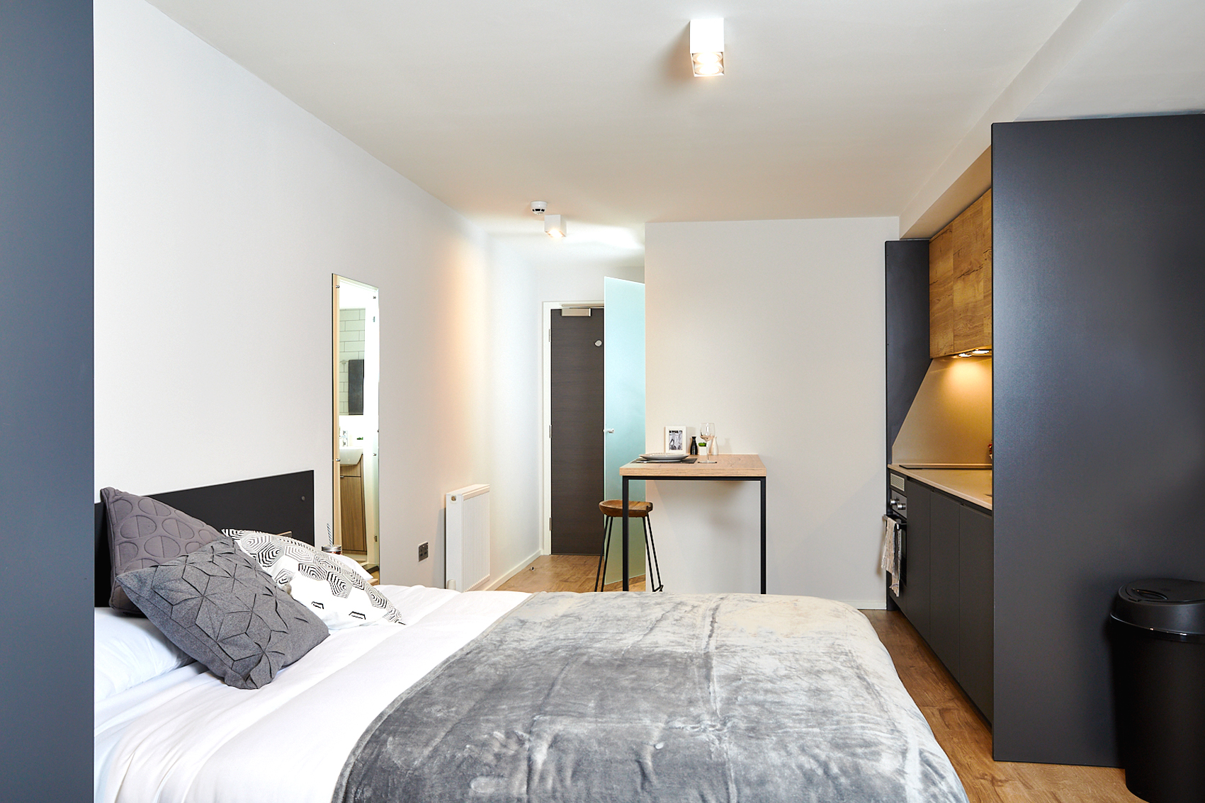 Located on Station Street, this modern studio apartment features a cozy bed with gray and white pillows, a small kitchenette with wood accents, and a bar stool by the high table. The visible bathroom entrance in the background complements its sleek, minimalist design.