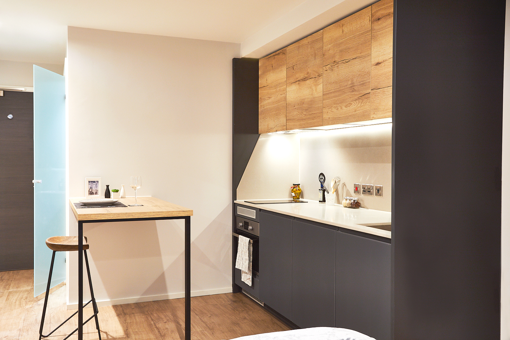 Nestled on Station Street, this modern, minimalist kitchen boasts wood and black cabinets, a compact sink, and an oven. A tall wooden table with a stool set for two features a wine glass and bubble decoration. The space is illuminated with warm lighting, creating an inviting atmosphere.