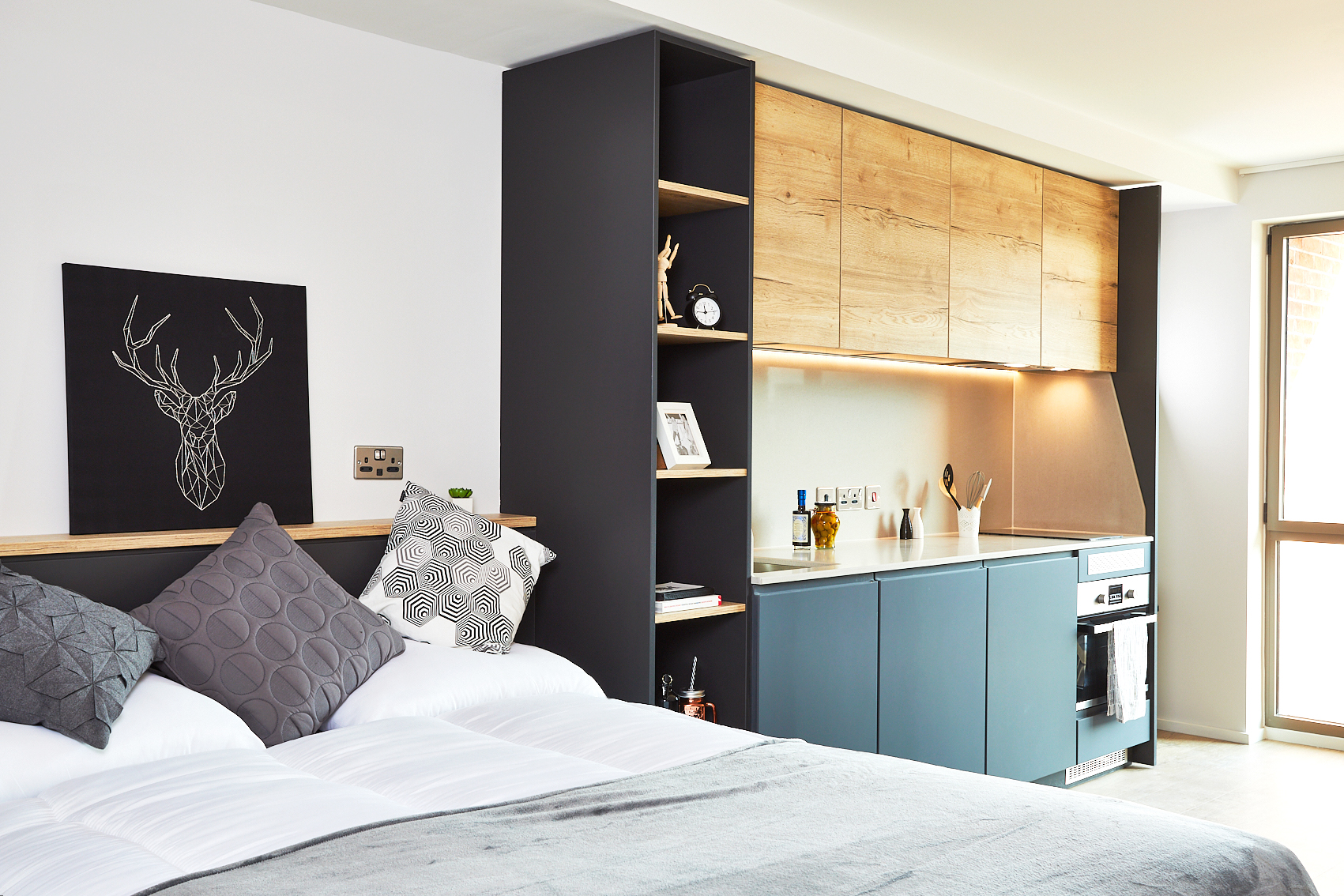 Discover this modern studio apartment on Station Street, featuring a bed with gray and patterned pillows. The kitchen area boasts blue cabinets, wooden shelves, and a sleek countertop. A black and white deer artwork enhances the wall next to the bed, all illuminated by abundant natural light from a large window.