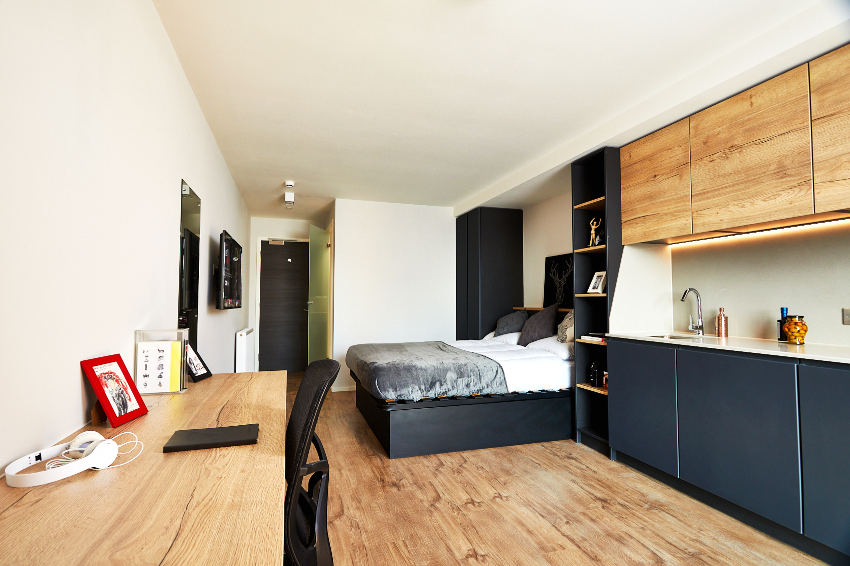 A modern studio apartment on Station Street featuring a bed with gray bedding, a wooden desk holding a laptop and headphones, and a kitchenette with wooden cabinets. The light wood flooring and neutral walls create a sleek, minimalist look.
