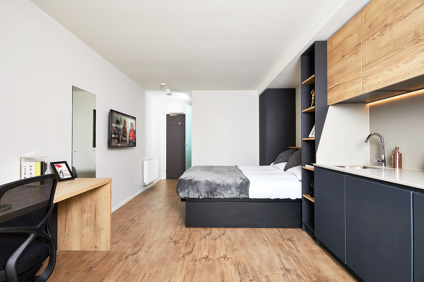Located on Station Street, this modern studio apartment features wooden flooring, a bed with gray bedding, integrated shelving, a kitchenette with sleek black cabinets, a desk and chair, a wall-mounted TV, and a mirror. The minimalist design is completed with a door at the far end.