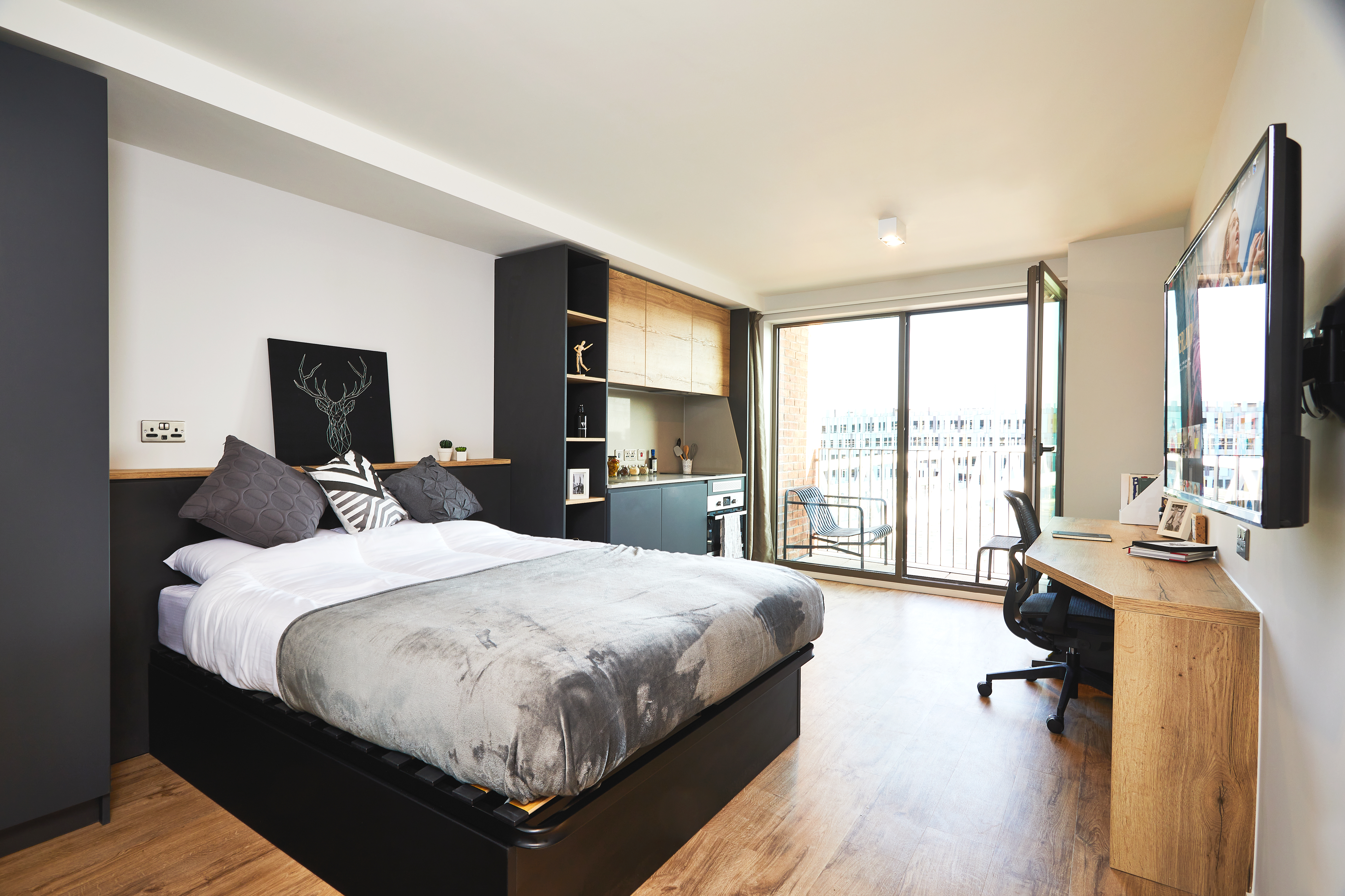 A modern, well-lit bedroom on Station Street features a double bed with gray and white bedding. A large TV is mounted on the wall opposite the bed. There's a desk and chair near sliding glass doors leading to a balcony, with cabinets and decorative items completing the space.