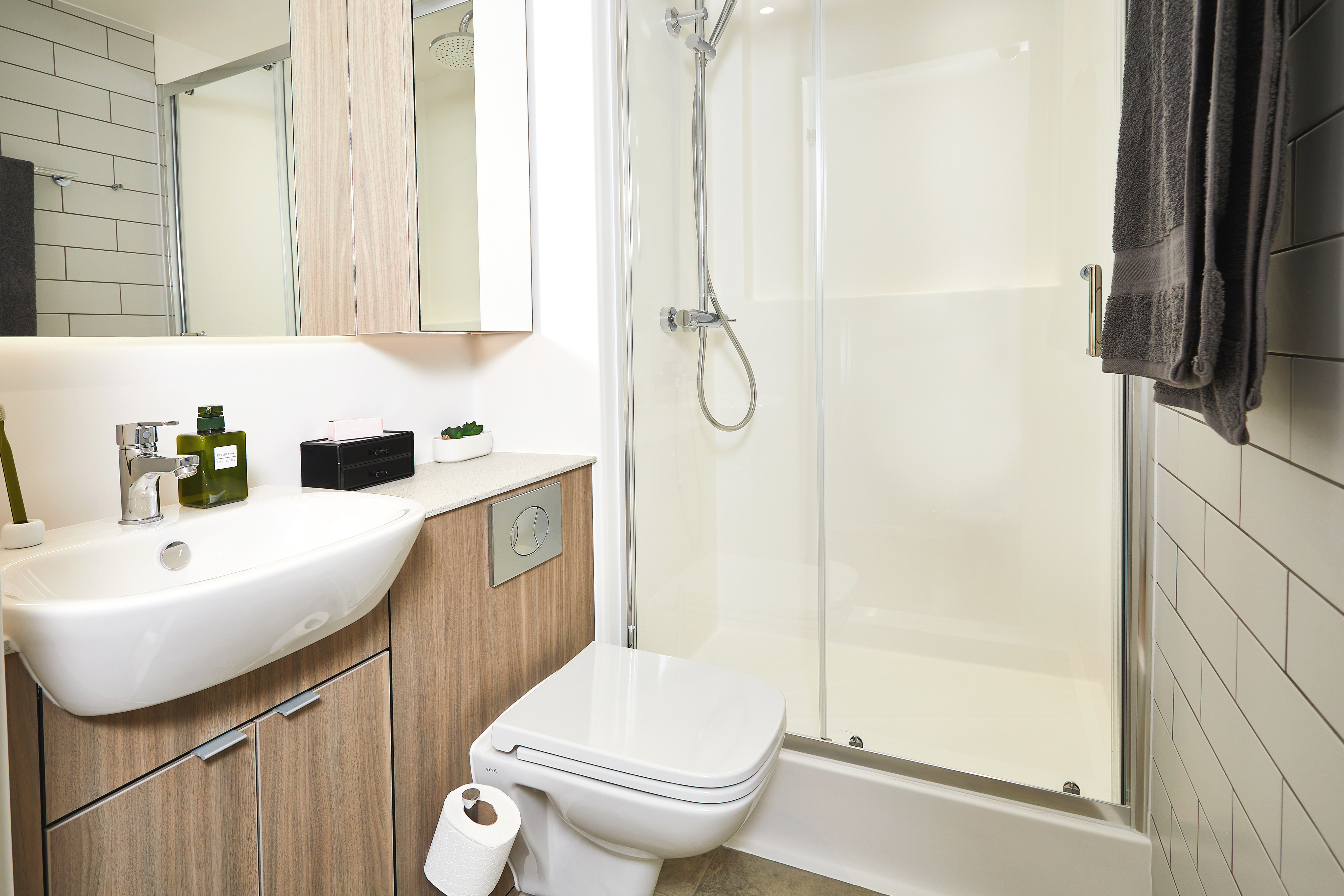Modern bathroom on Station Street, featuring a glass-enclosed shower, a wooden cabinet with a white sink, and a wall-mounted toilet. A towel hangs gracefully on the glass door beside a visible toilet paper roll. The minimalist decor is completed with light-toned tiles for an elegant finish.