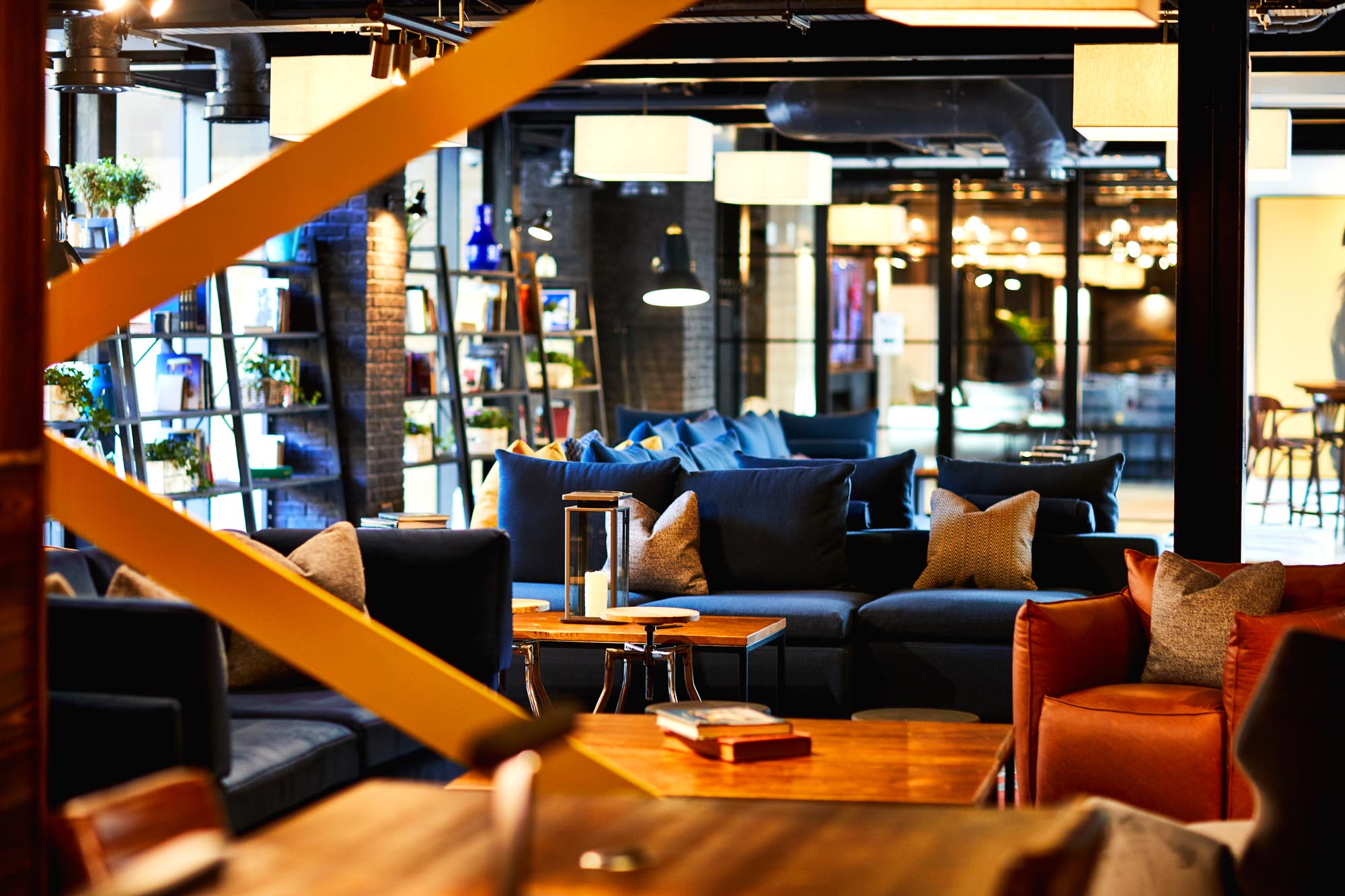 A cozy lounge in Fountainbridge with modern furniture, featuring blue and brown couches, wooden tables, and decorative shelves with books and plants. Soft lighting creates a warm ambiance, while large windows allow natural light to filter in.
