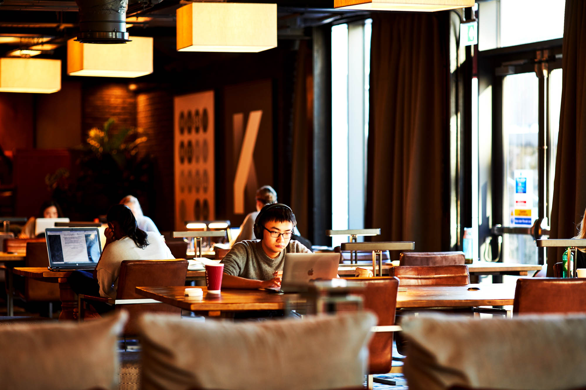 A cozy cafe in Fountainbridge, with warm lighting showing people seated at wooden tables, working on laptops. A person in the foreground focuses on their screen, with coffee cups nearby. Large windows allow natural light to filter in.