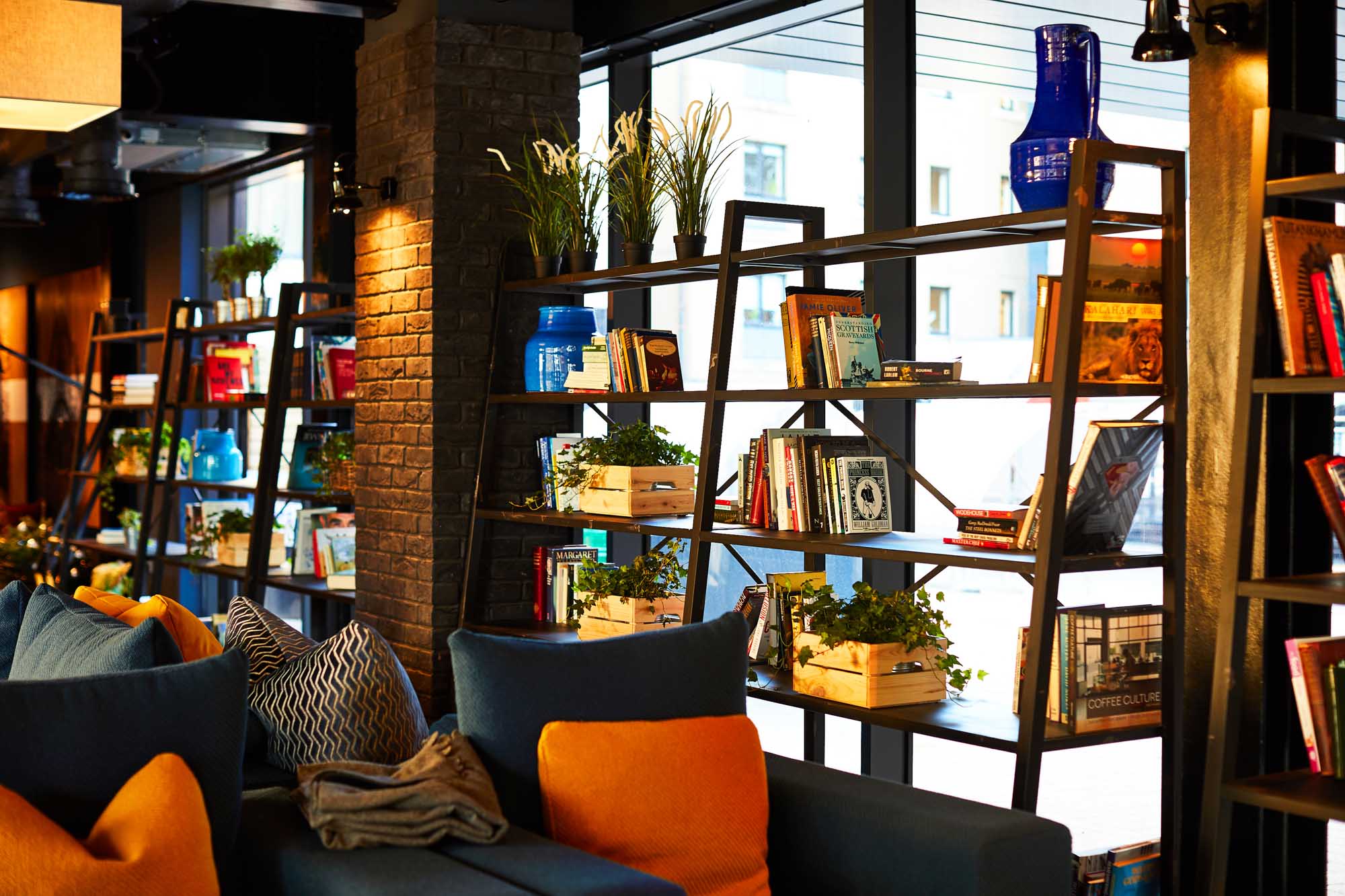 Modern interior in Fountainbridge, boasting bookshelves filled with books, potted plants, and blue vases. Shelves line large windows that offer an urban view. Cozy seating with blue and orange cushions sits in the foreground, creating a warm, inviting atmosphere bathed in natural light.