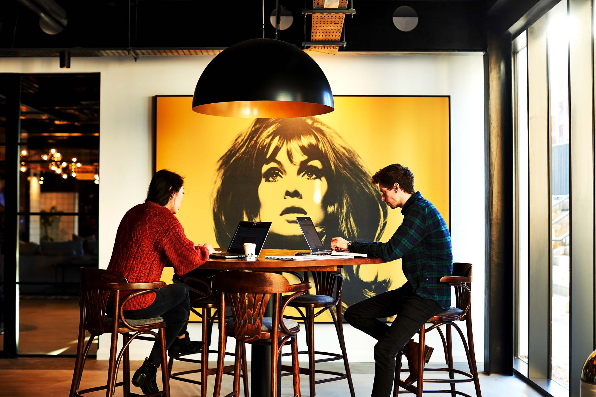Two people working on laptops at a high table in a modern space in Fountainbridge, with a large, yellow-toned portrait of a woman on the wall behind them. The area is lit by an elegant black dome light fixture.