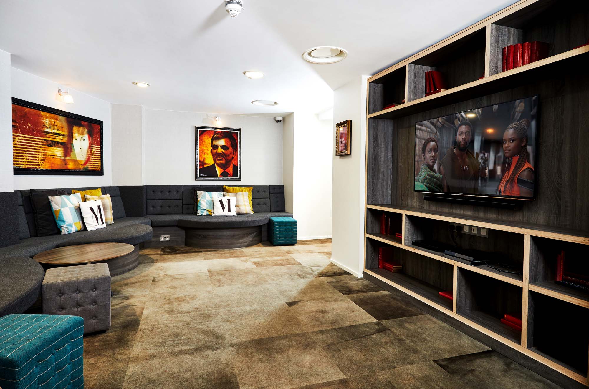 A modern lounge in Portland House features circular seating, colorful pillows, and a large flat-screen TV displaying a movie scene. The room is adorned with artwork on the walls, bookshelves, and a carpet showcasing different shades of brown.
