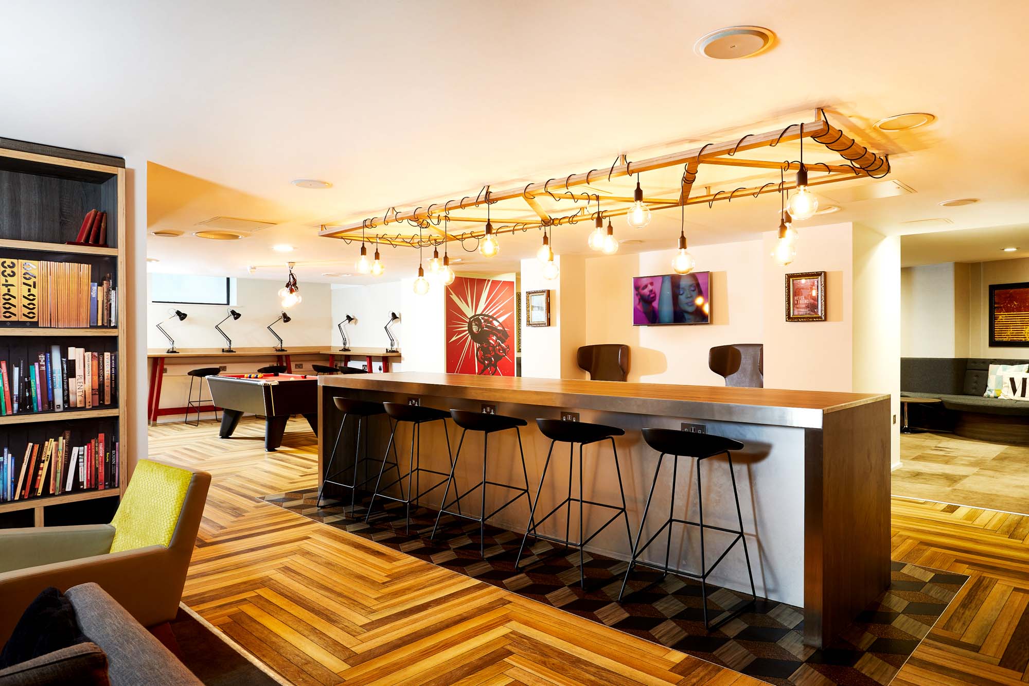 The Portland House offers a modern lounge area with a long bar table and four black stools. Exposed bulbs hang above, illuminating the wooden floor, bookshelves, and colorful artwork. A seating area with tables and chairs is beautifully visible in the background.