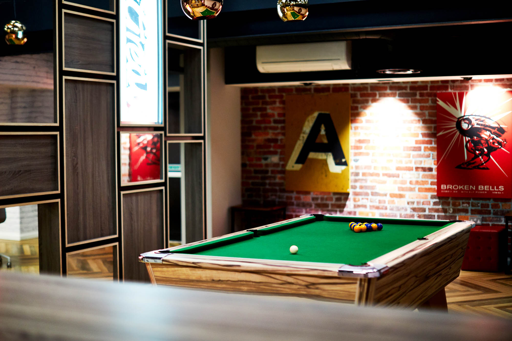 A cozy game room featuring a pool table with green felt, surrounded by modern wood-paneled walls. A brick wall in the background displays colorful artwork, including the letter "A." Soft lighting creates a relaxed atmosphere.