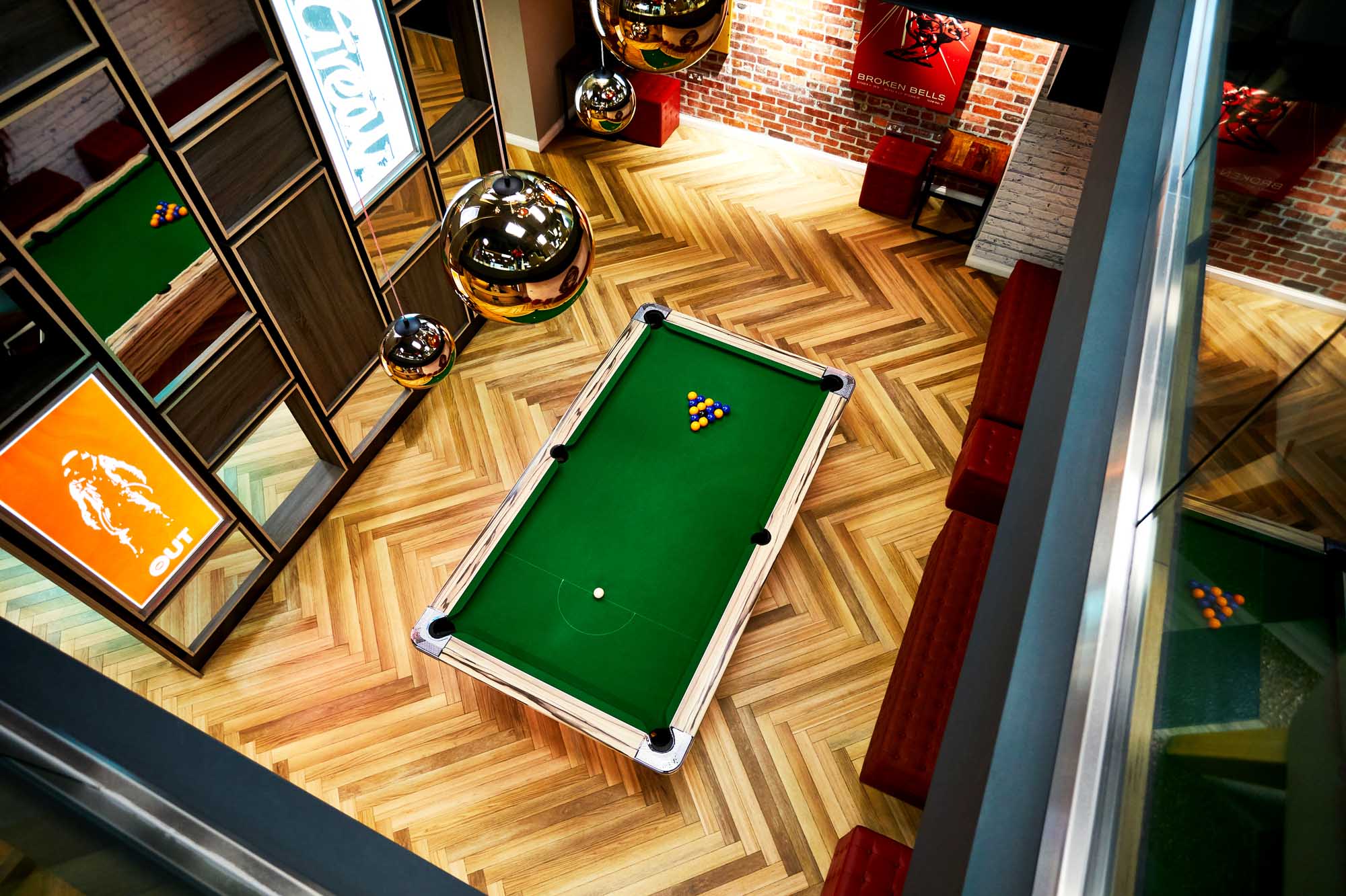 Overhead view of a stylish room in the house featuring a green-felt pool table at its center. The space boasts parquet flooring, brick walls, wall shelves, golden spherical lights, and framed artworks. Pool balls are arranged in a triangle on the table.