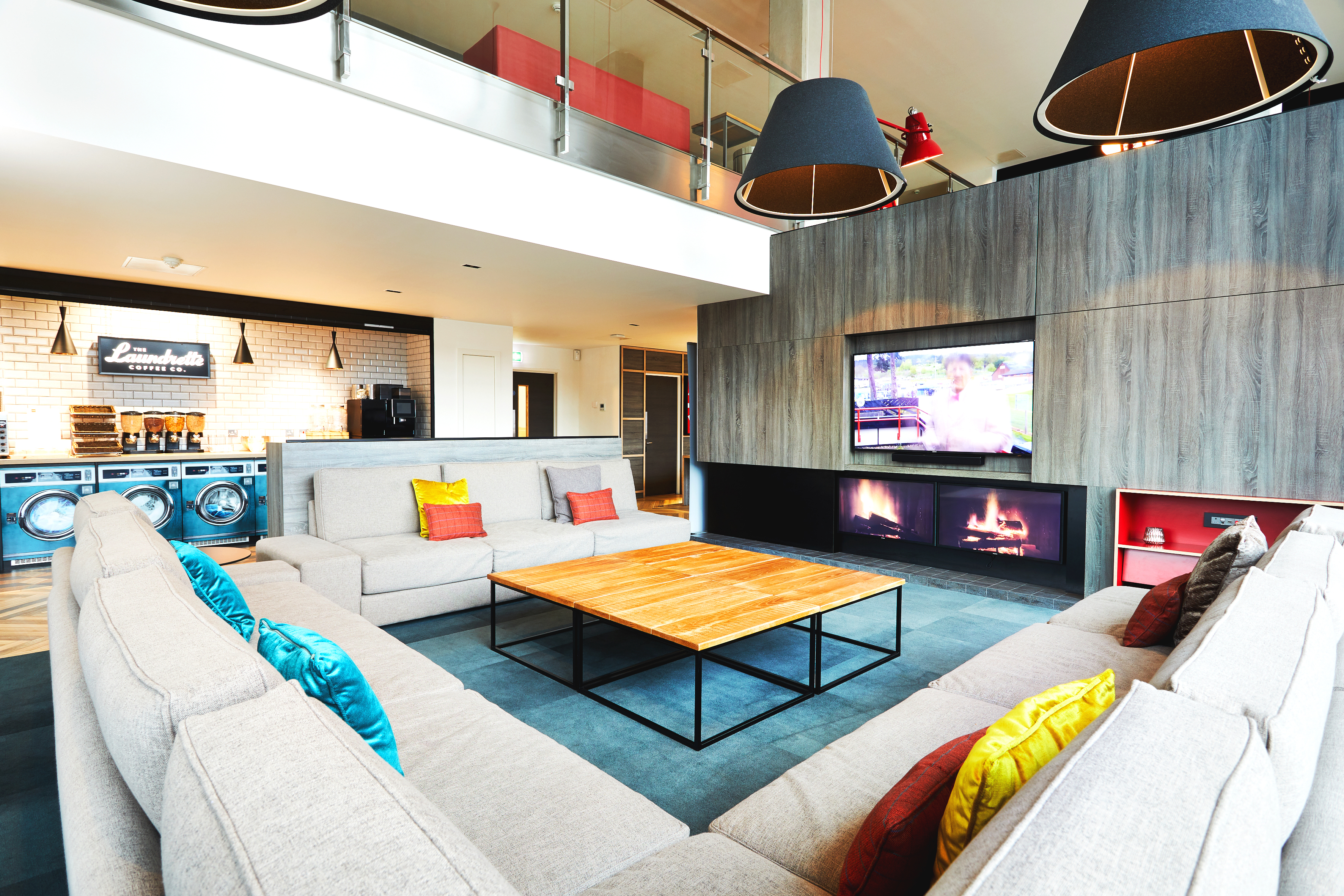 A modern living room with gray sofas forming a U-shape around a wooden coffee table. A wall-mounted TV and fireplace are on the right. The room includes stylish black pendant lights and an open laundry area with blue machines and a sign.