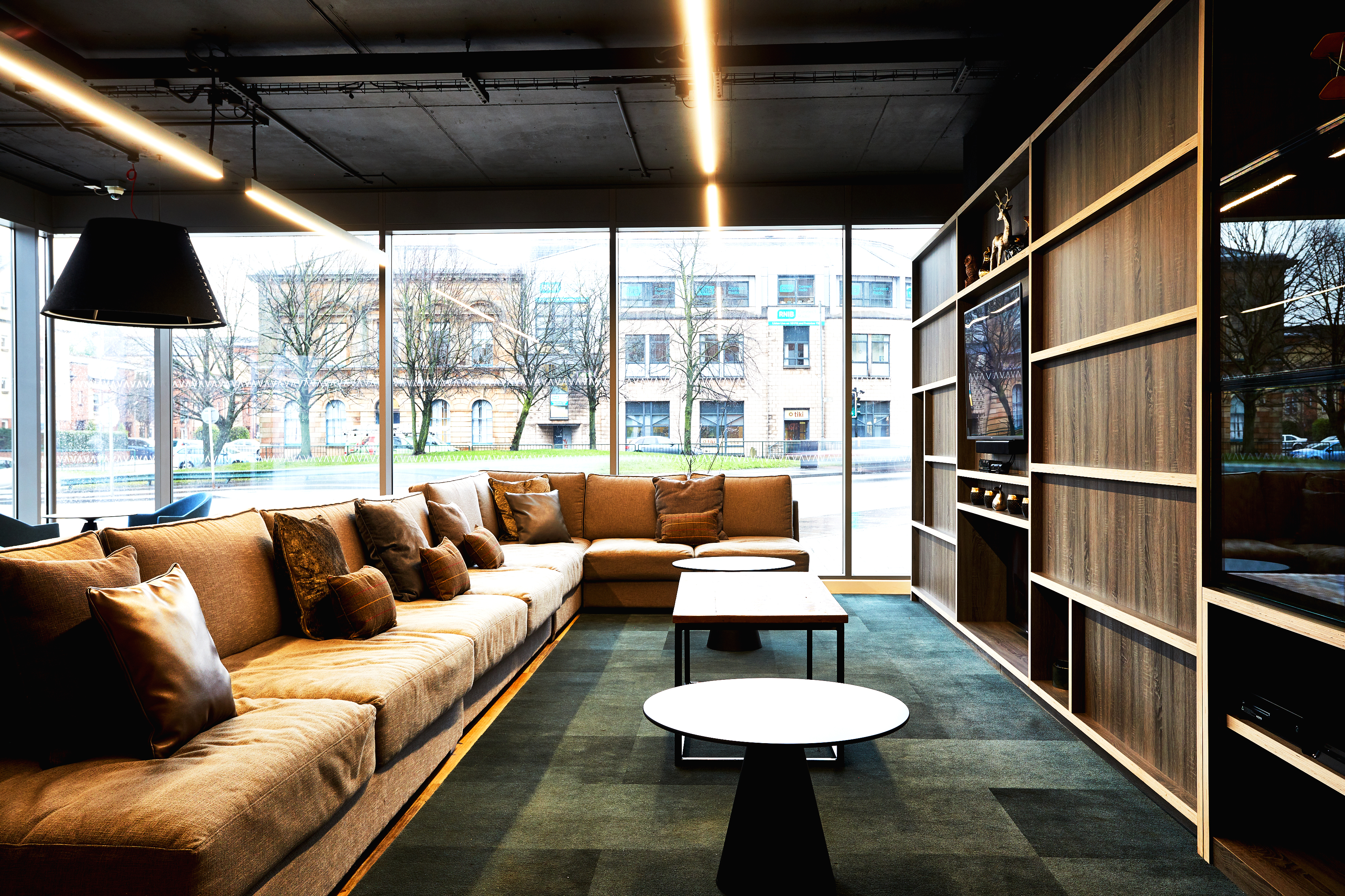 Modern lounge with large brown sectional sofa, decorative pillows, and three small round tables. Floor-to-ceiling windows offer a view of an urban street with buildings and trees. Wooden shelving on the right and linear ceiling lights above.