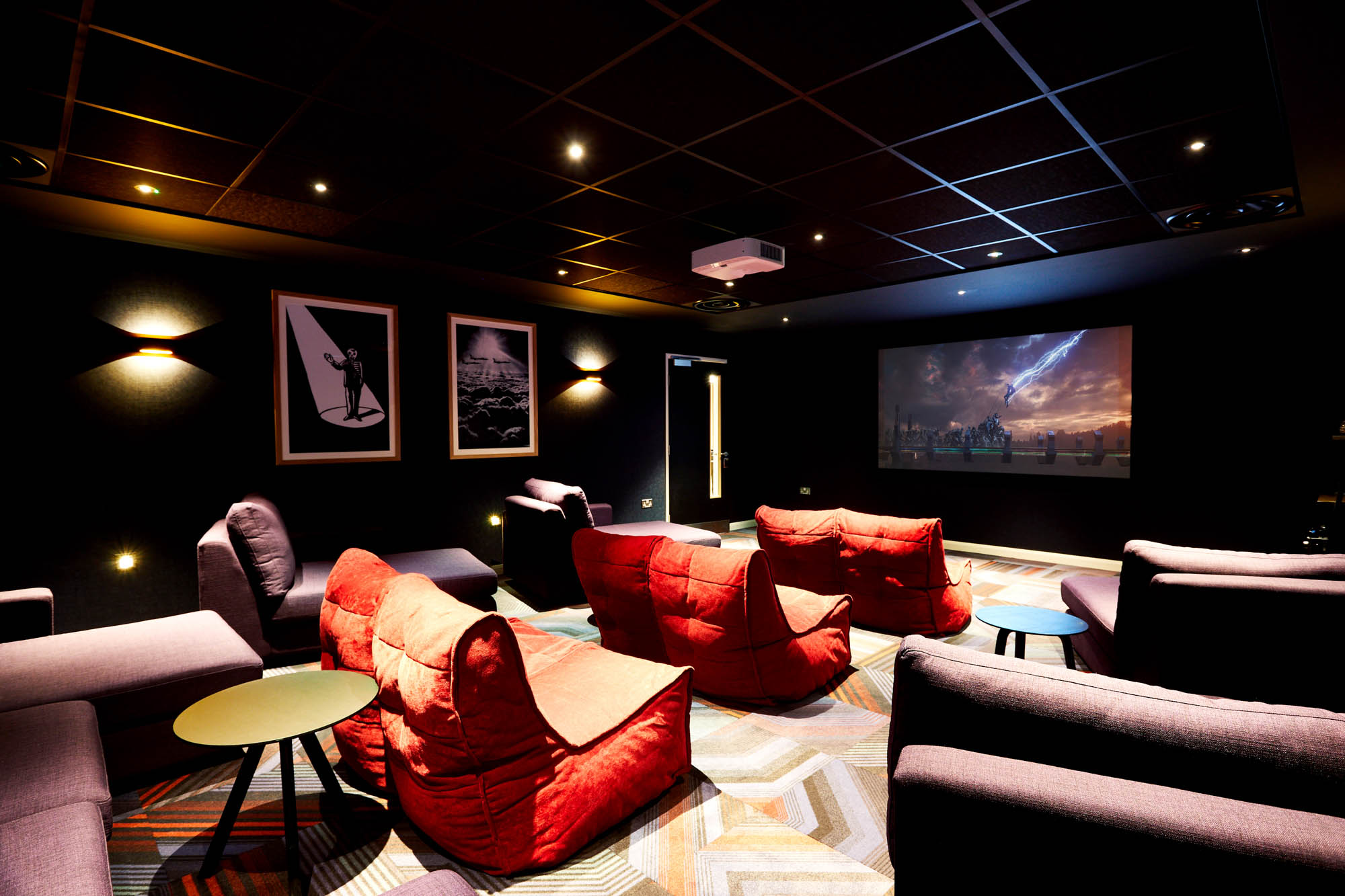 A cozy home theater with dim lighting and plush red beanbag chairs is artfully arranged in a circular formation facing the large projection screen. The room's dark walls are adorned with black and white art, while a patterned carpet adds warmth to this modern setting.