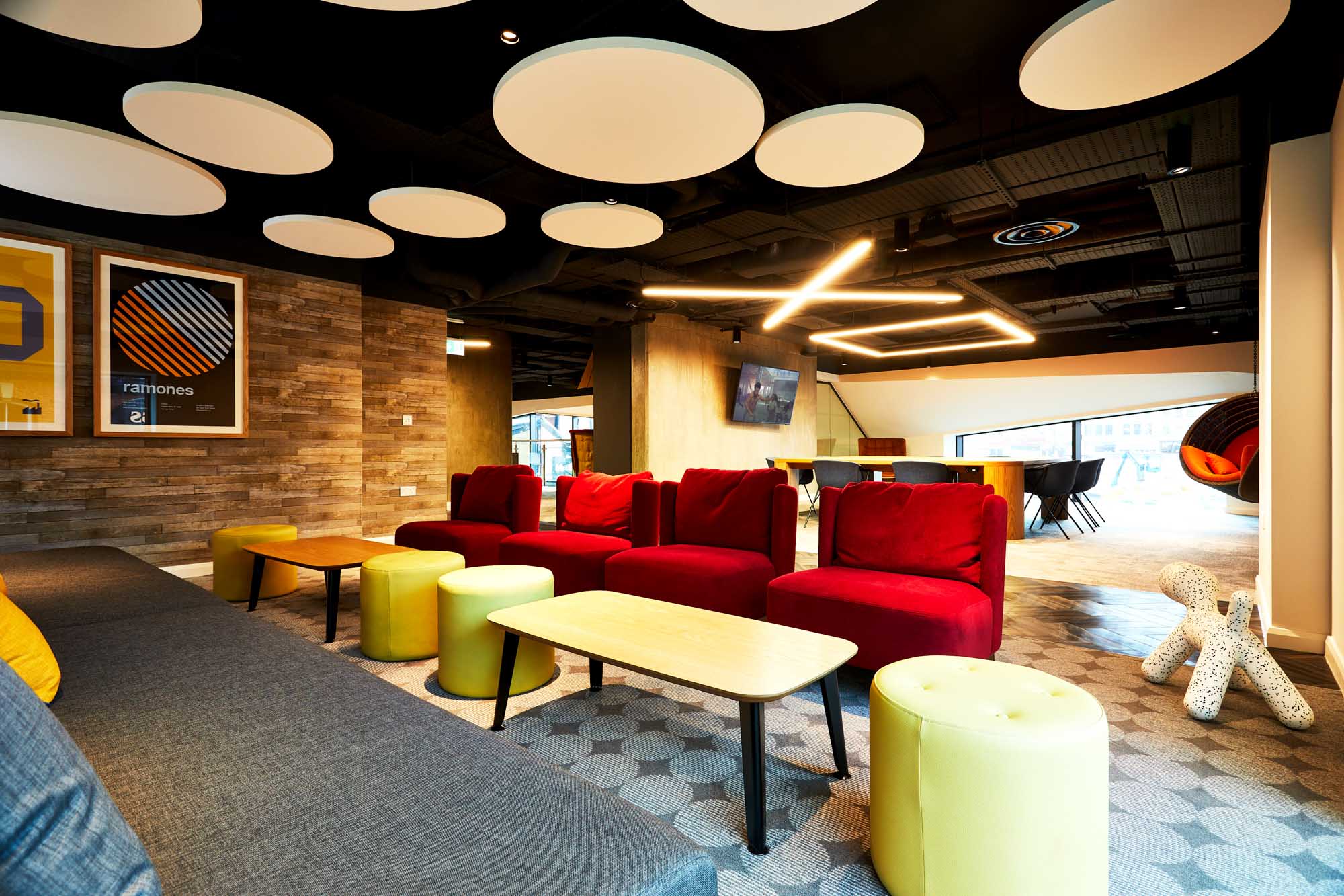 Modern lounge with red and yellow seating, wooden tables, and circular ceiling panels. Geometric lights hang above, casting patterns of circles and squares, while artwork decorates the walls. The room features a mix of natural light and cozy ambiance.