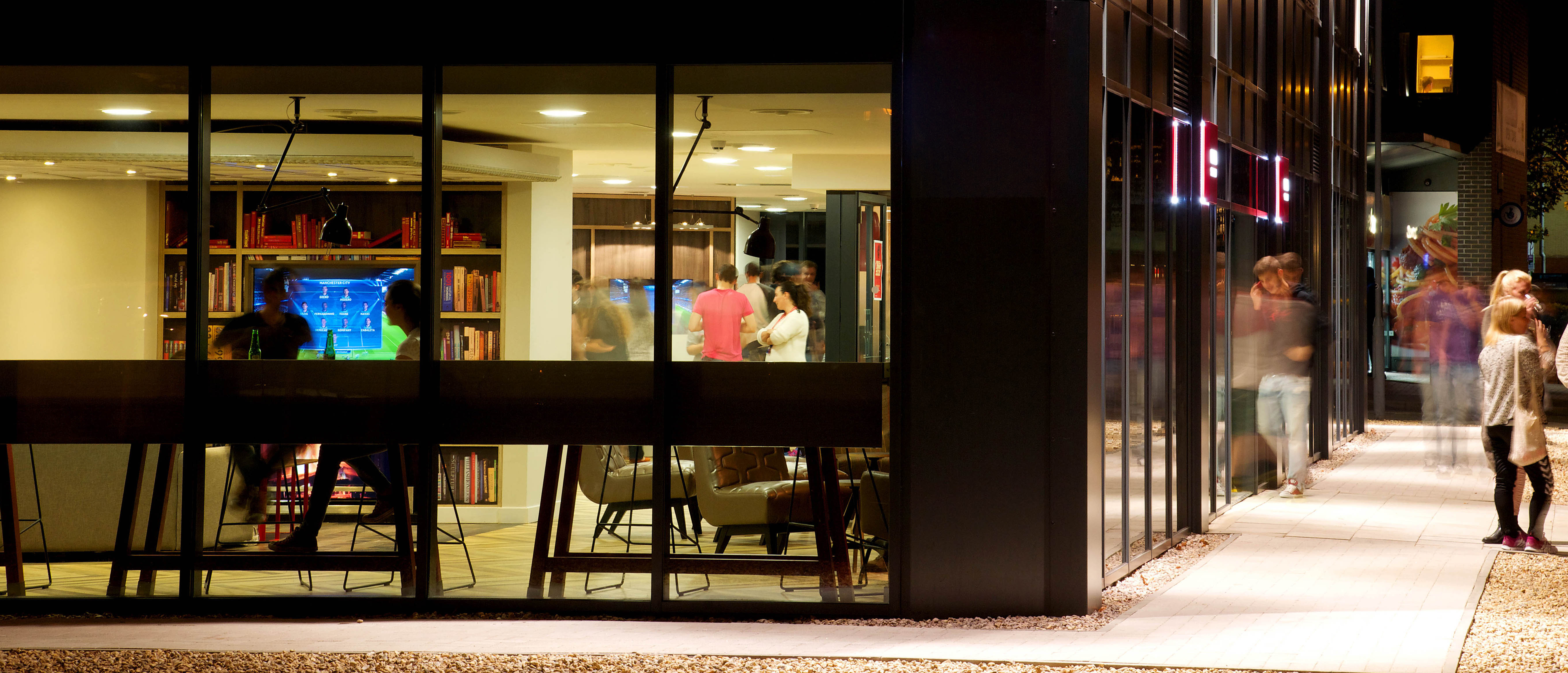 A modern building with large glass windows showcases a brightly lit interior, where visitors engage with interactive digital displays. Outside, the blurred motion of people walking along the bustling sidewalk adds to the dynamic scene.