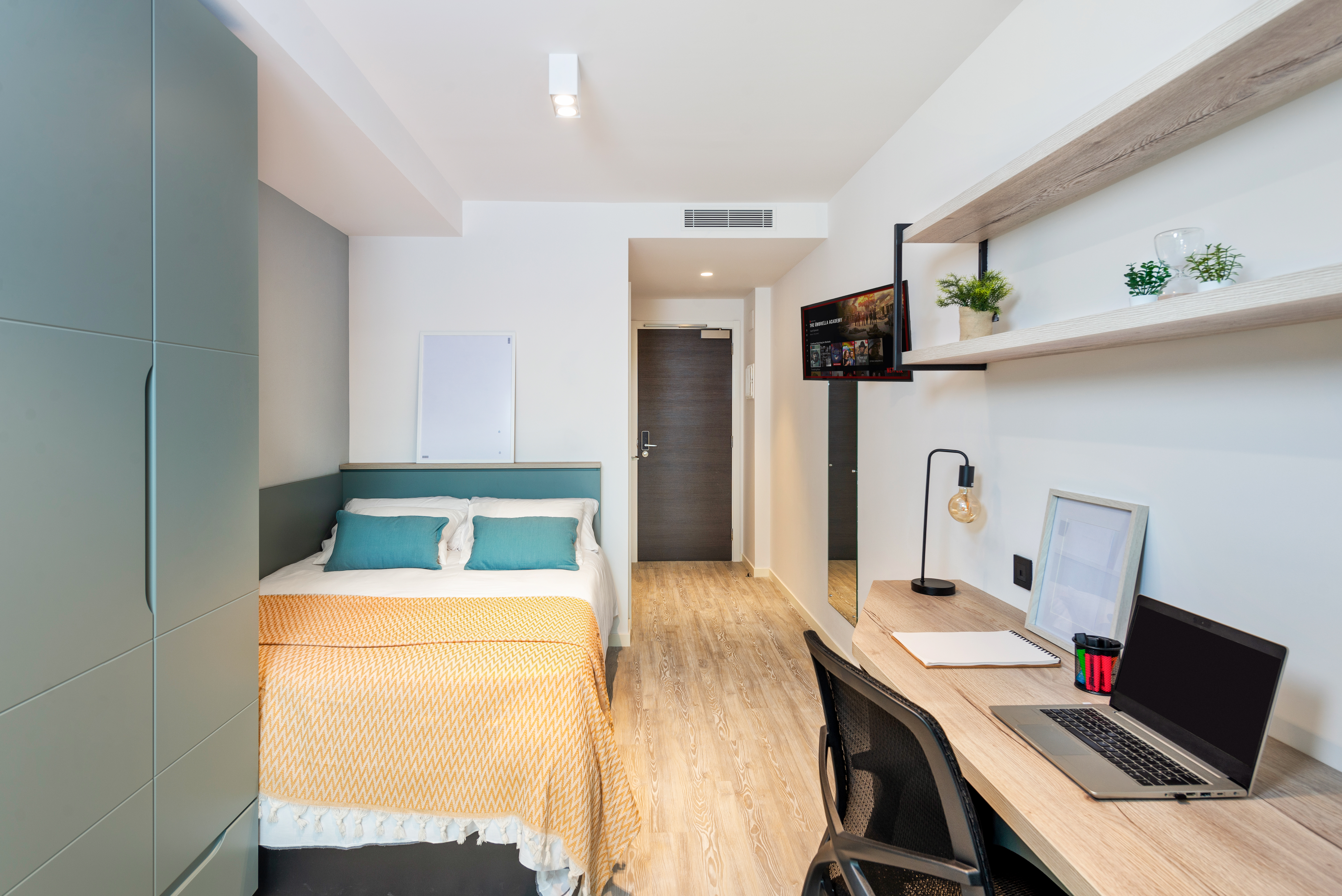 A modern, minimalist bedroom features a bed with turquoise pillows and an orange blanket. There is a wooden desk with a laptop and a desk lamp, and shelves with plants above. A flat-screen TV is mounted on the wall near the door.