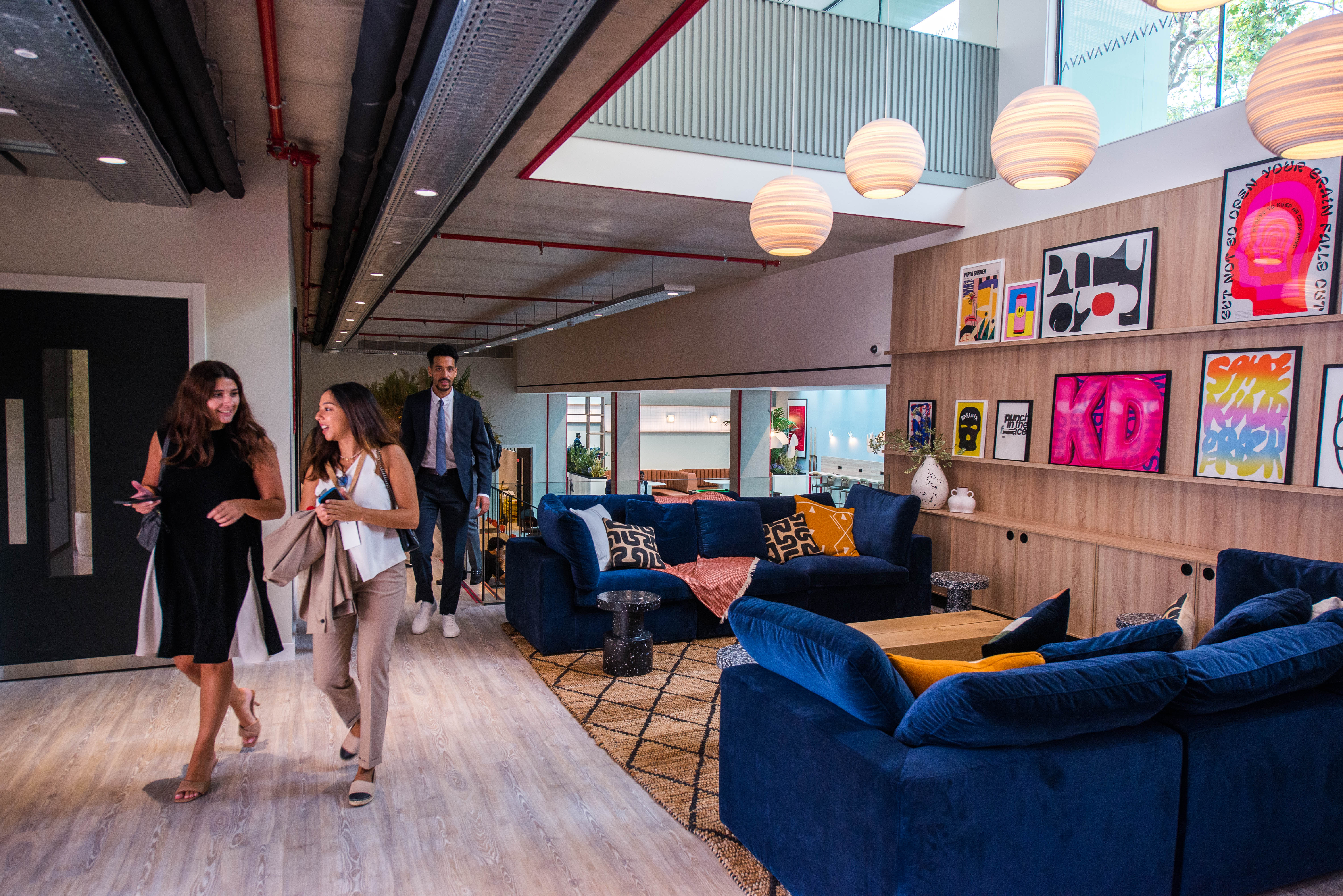 A modern office space in the UK features two women and a man walking and talking. The interior boasts a cozy lounge area with blue sofas, colorful artwork, and round hanging lights. Large windows let in natural light, creating an inviting environment for professionals.