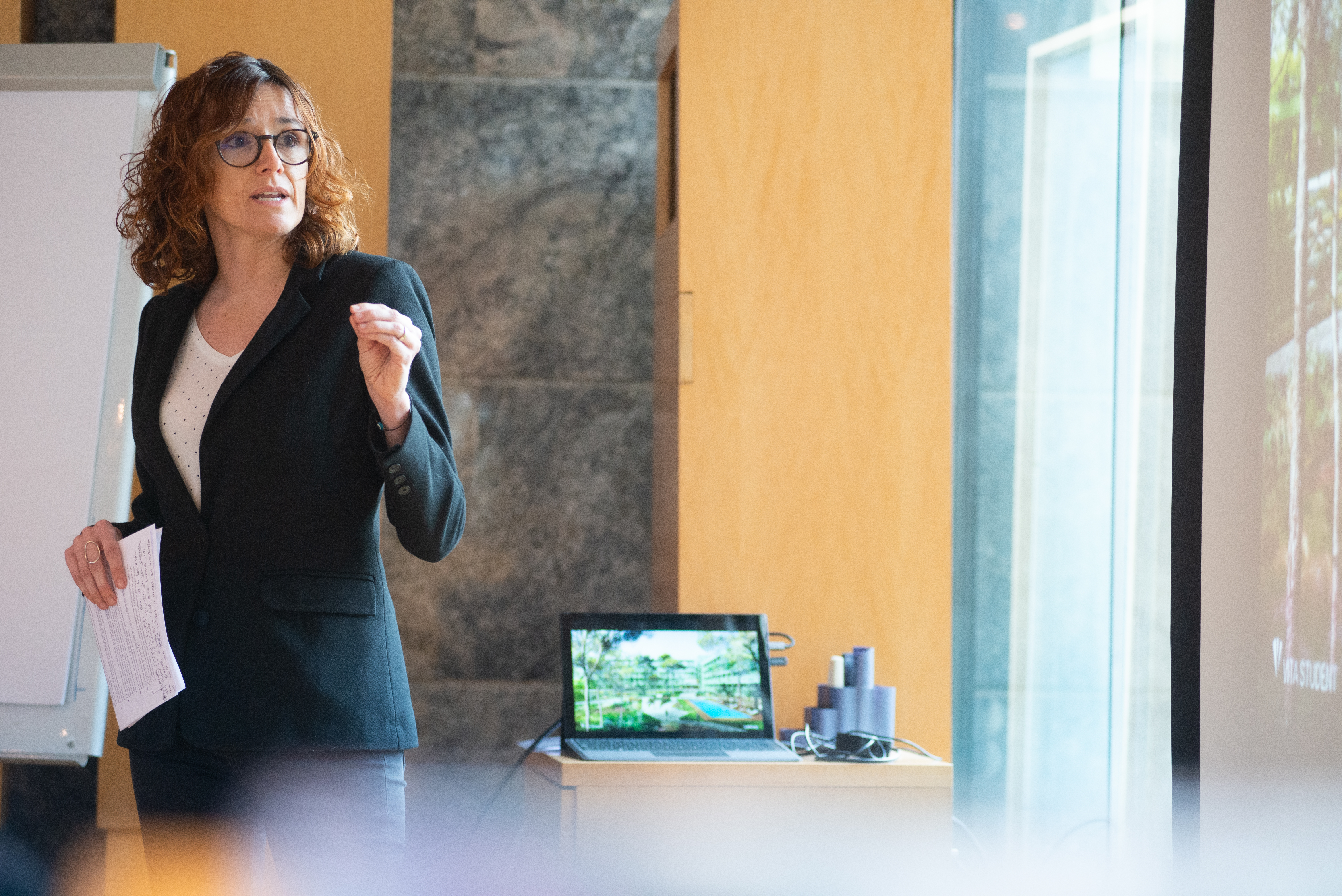 A woman in a black blazer and glasses is giving a presentation on start-up support. She's standing next to a screen, speaking, and holding a remote. A laptop displaying an image is on a table behind her, with a whiteboard in the background.