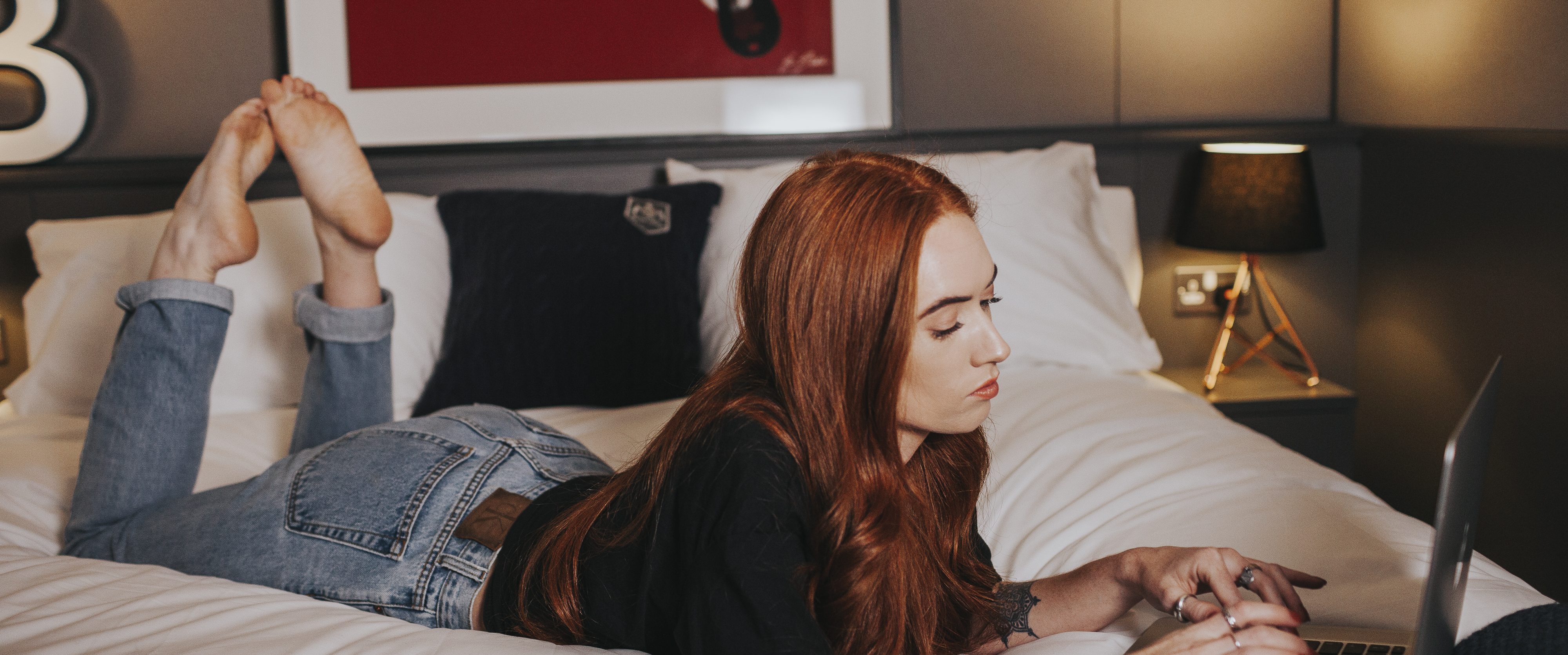 A person with long red hair is lying face down on a bed, working on a laptop, exploring new learning strategies. They are wearing a black top and blue jeans in a room with modern decor, featuring a framed picture on the wall and a lamp on the nightstand.