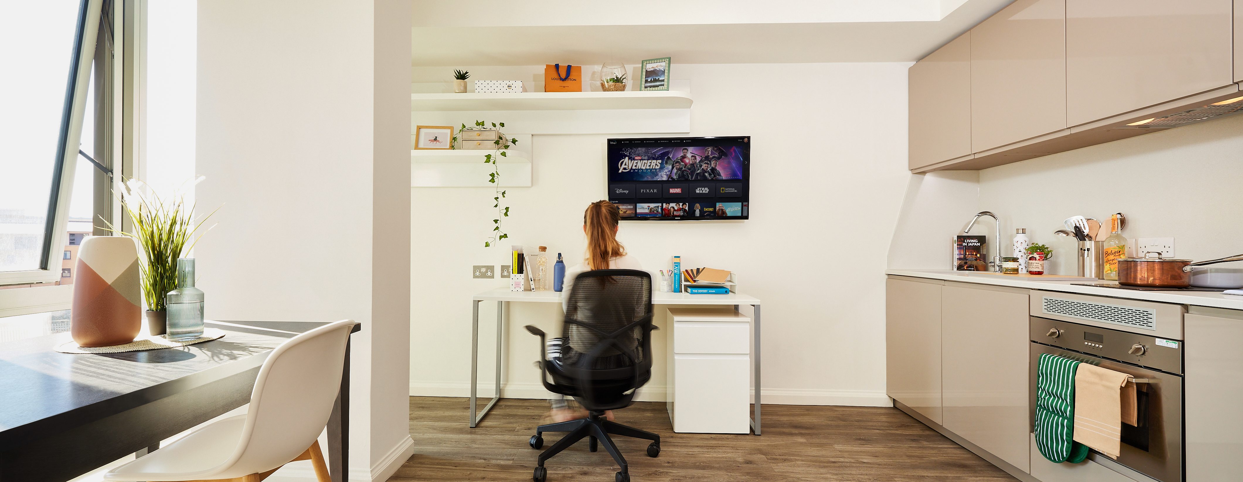 A woman sits at a desk in a modern apartment, utilizing time management tips as she watches a TV mounted on the wall. The room features a kitchen with open cabinets and a dining table by the window, adorned with plants and minimalistic decor, ideal for students balancing studies and relaxation.