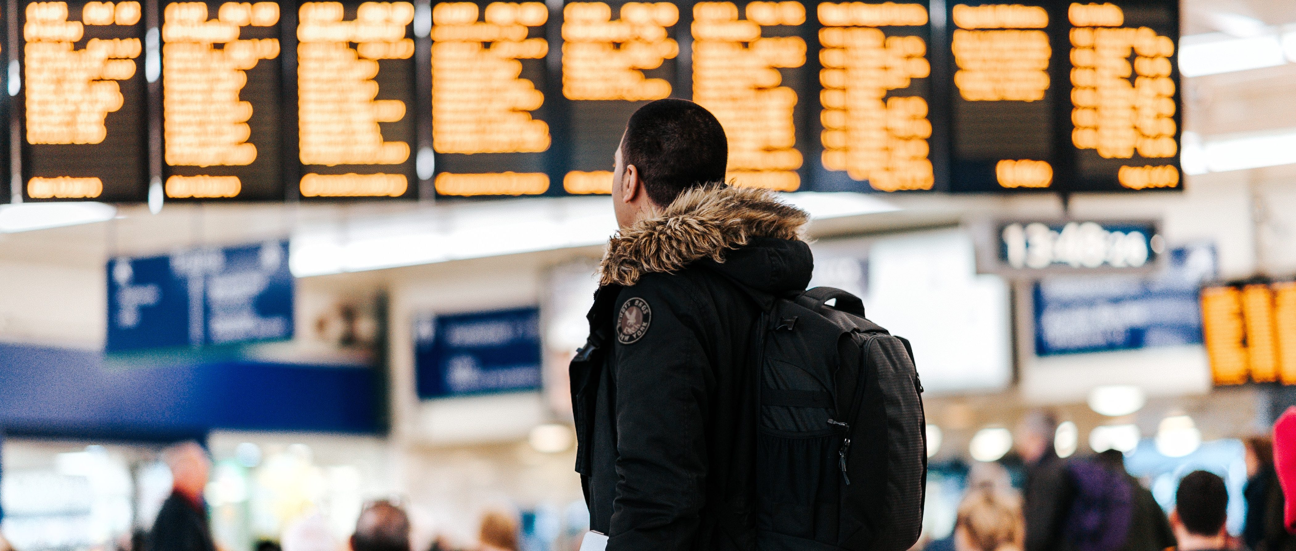 A person with a backpack looks up at a large, brightly lit digital departures board displaying numerous flights in a busy station, contemplating the excitement of studying abroad. The setting is bustling with people and signs, each carrying their own stories and destinations.
