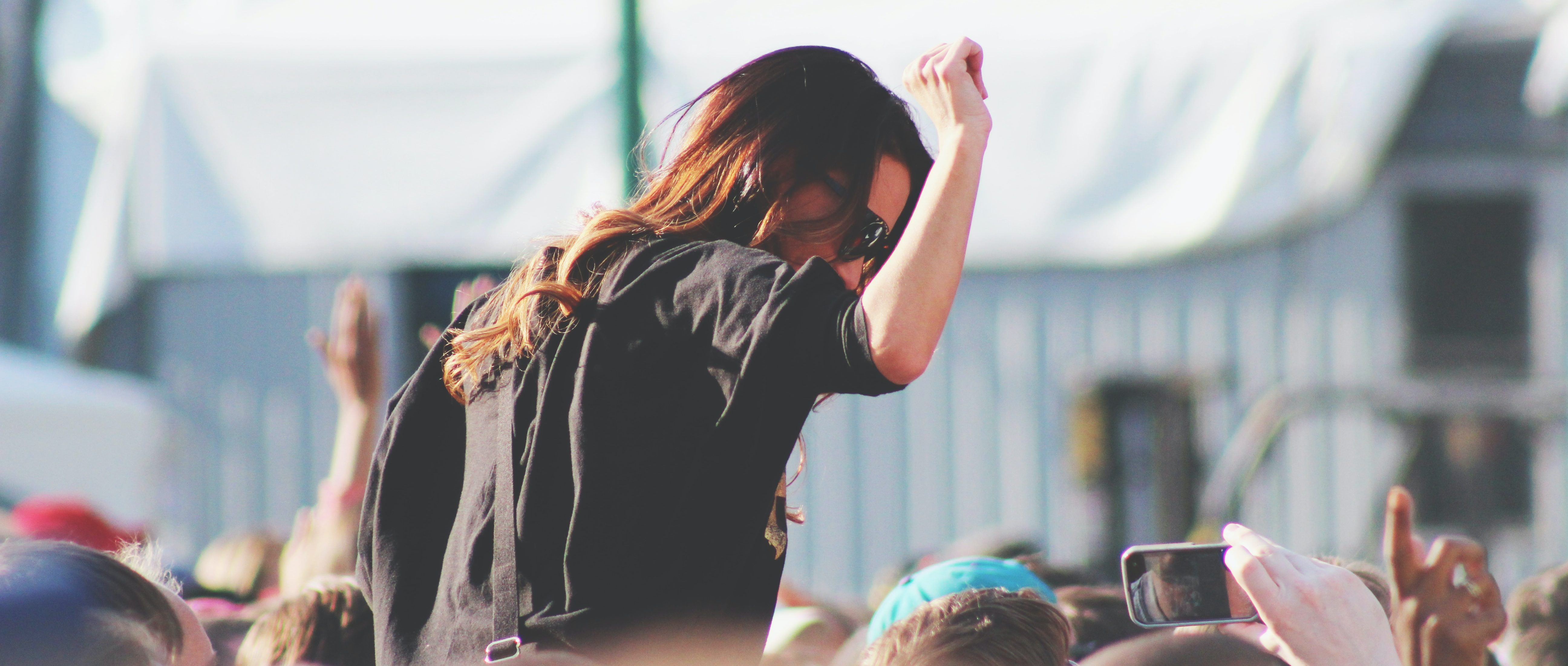 At the lively Parklife Festival, a person with long hair and sunglasses, dressed in a black shirt, is perched on someone's shoulders. With one arm raised high under the summer sun, they join a sea of phones capturing the vibrant moment of music and joy.