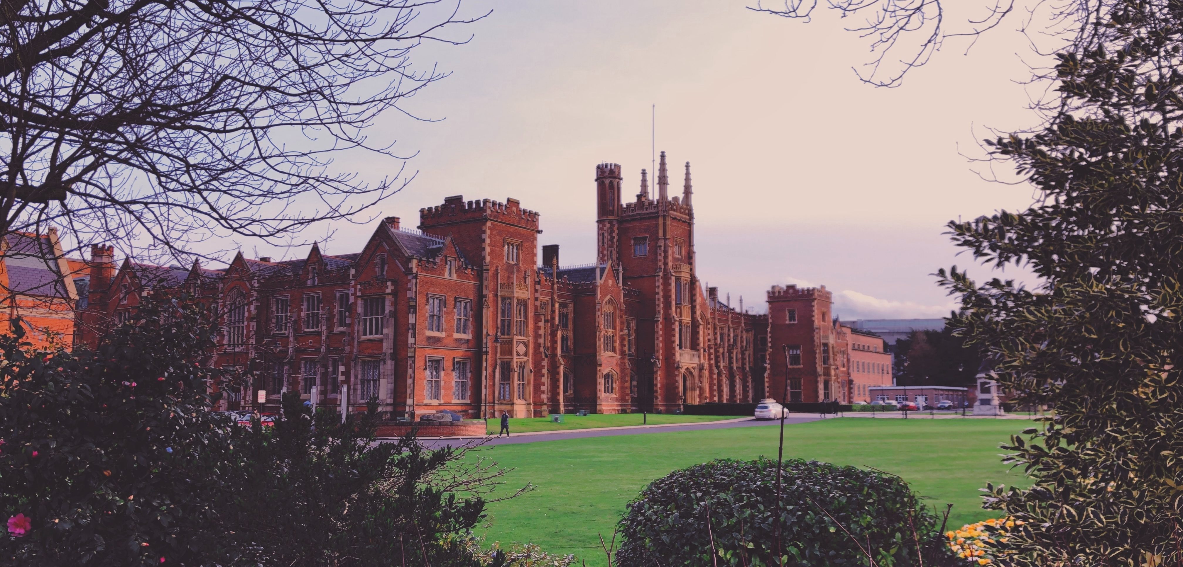 A historic red-brick building with tall towers stands on a lush green lawn in Belfast. Framed by trees under an overcast sky, this iconic site is one of the city's must-visit attractions, offering a glimpse into its rich past and a perfect addition to your list of things to do.