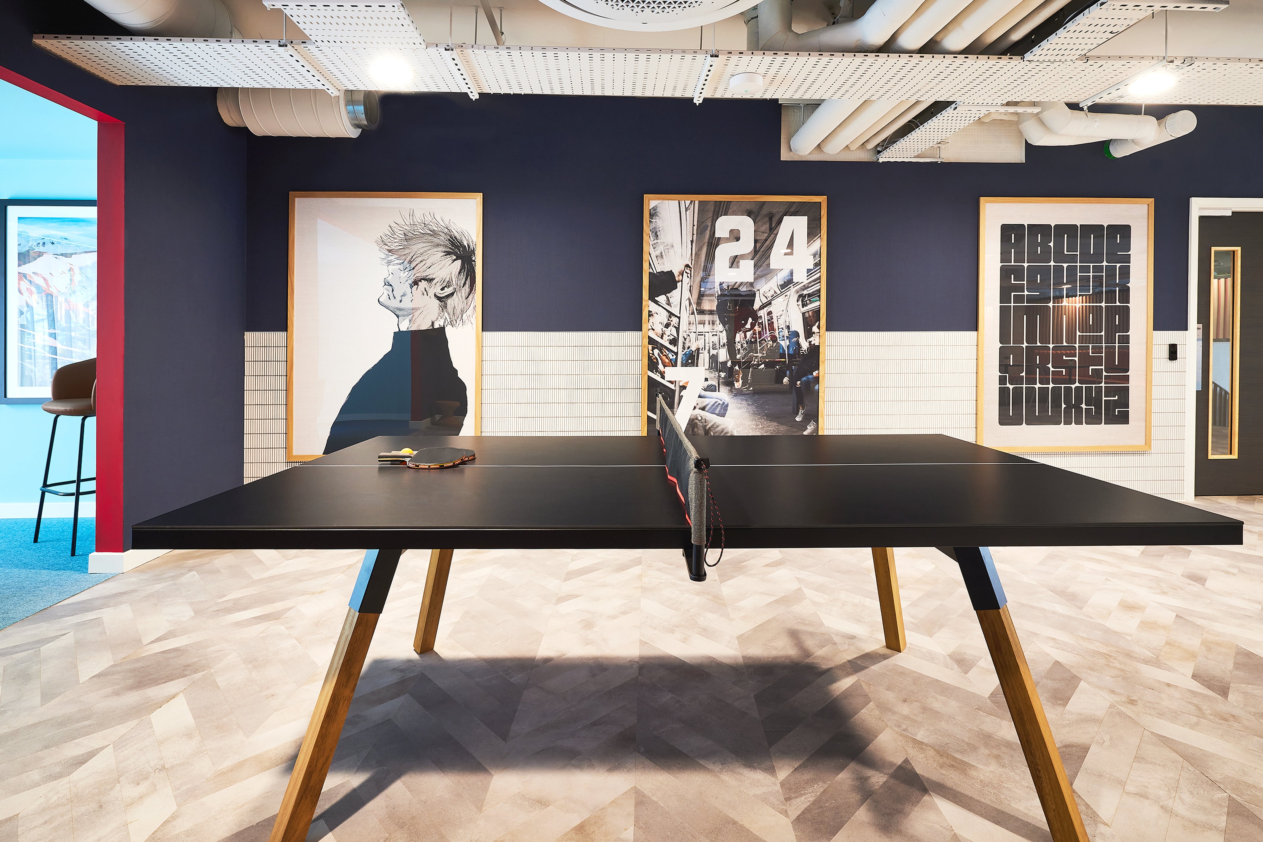 A modern room in India showcases a table tennis setup with artwork adorning the walls, a dark blue upper wall, and a white tiled lower section. The light herringbone patterned floor leads to an open doorway where a stool is visible, creating an ideal spot for relaxing or studying.