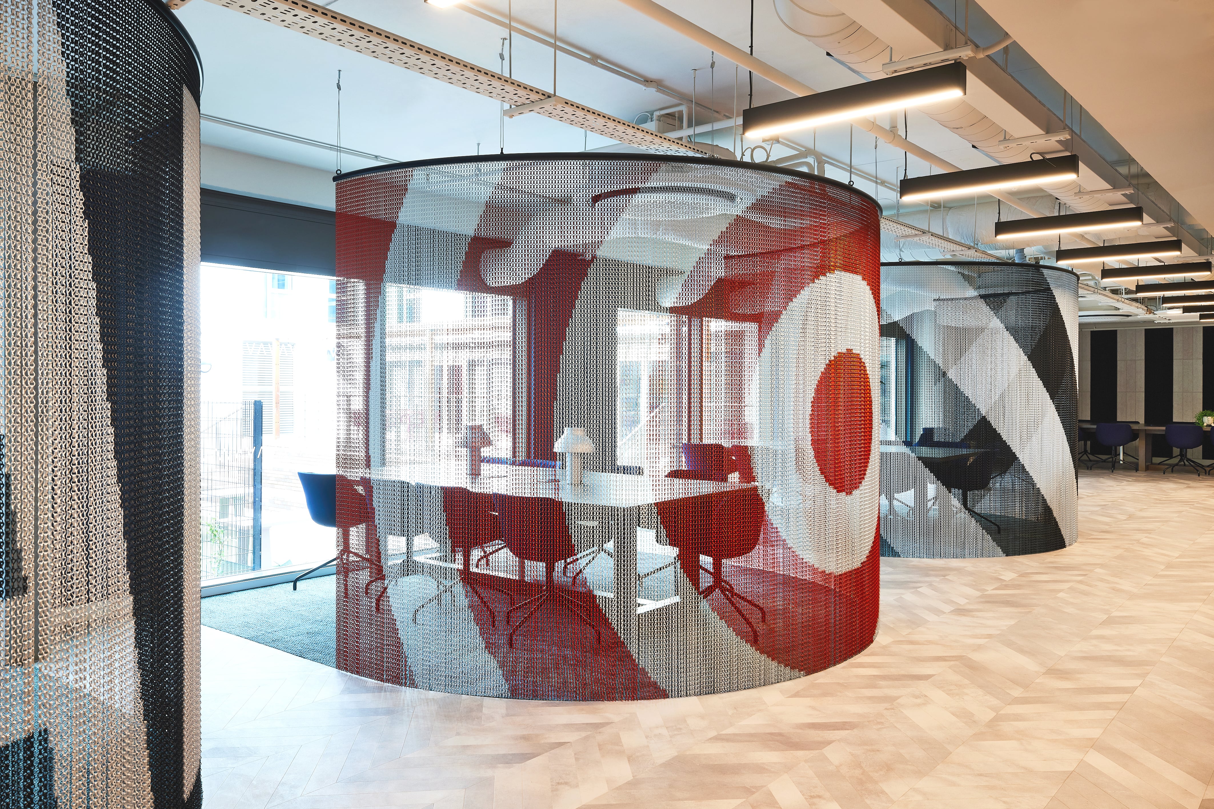 A modern office space reminiscent of India's vibrant aesthetics, featuring circular meeting areas enclosed by transparent, multicolored mesh curtains. The design boasts bold patterns in red, gray, and white. It's a perfect setting for studying or work with ample natural light from large windows and linear overhead lights.
