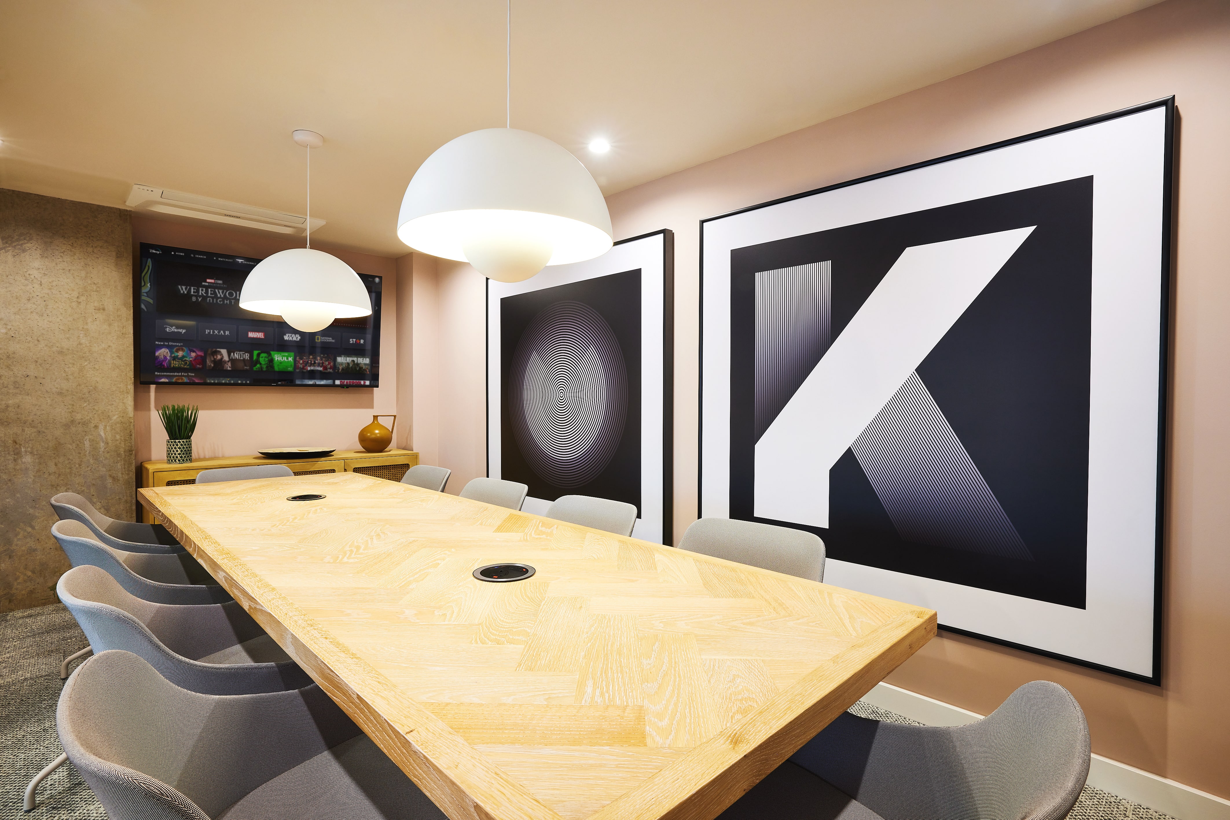 Modern conference room with a long wooden table surrounded by gray chairs, perfect for studying. Two large abstract artworks hang on the wall, their colors reminiscent of India. Pendant lights hang from the ceiling, and a TV is mounted in the corner above a console table.