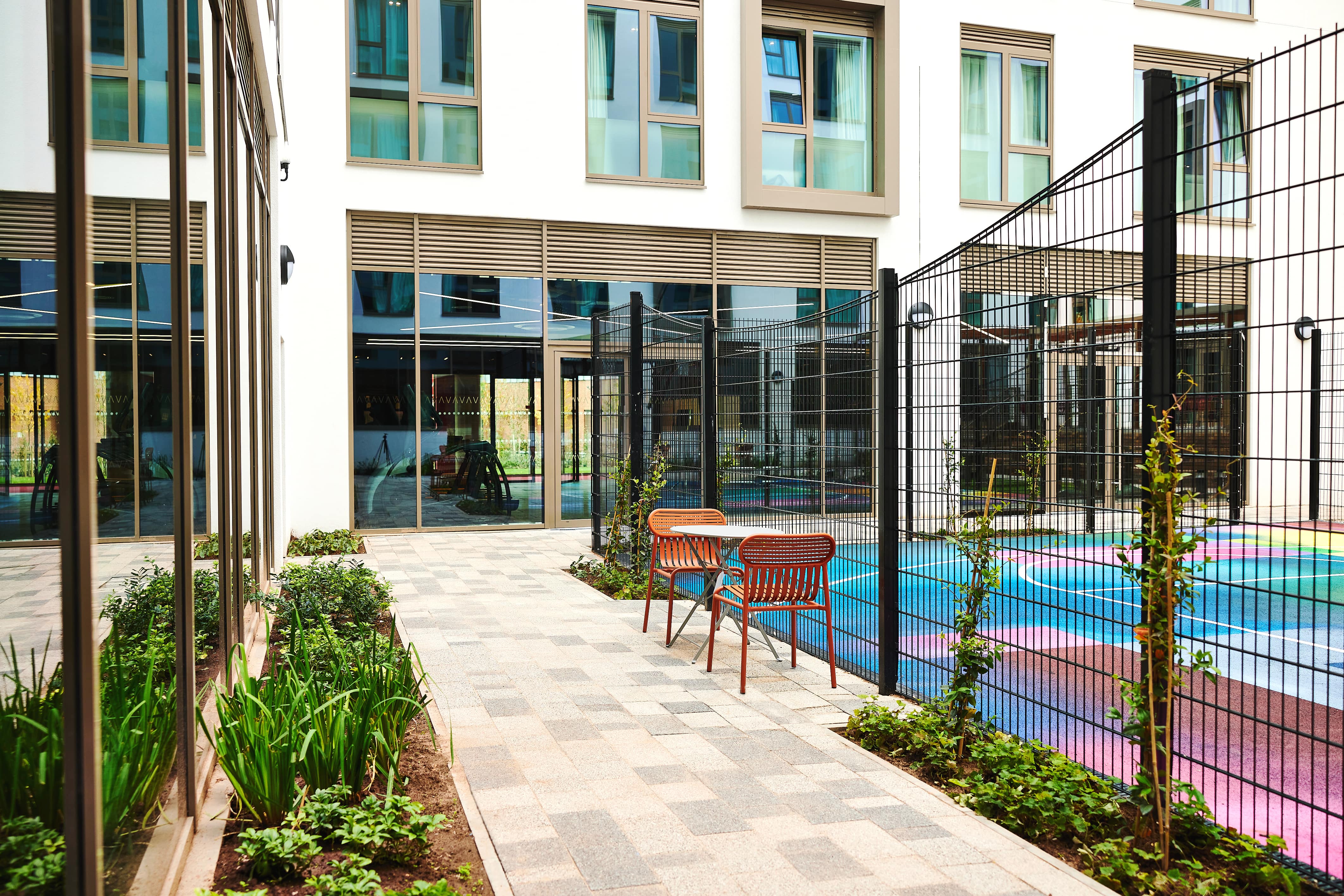The courtyard, reminiscent of a tranquil study spot in India, boasts a paved walkway flanked by plants and a wooden table with chairs. Colorful outdoor seating peeks through a black fence, while the building's large windows showcase its modern architectural style.