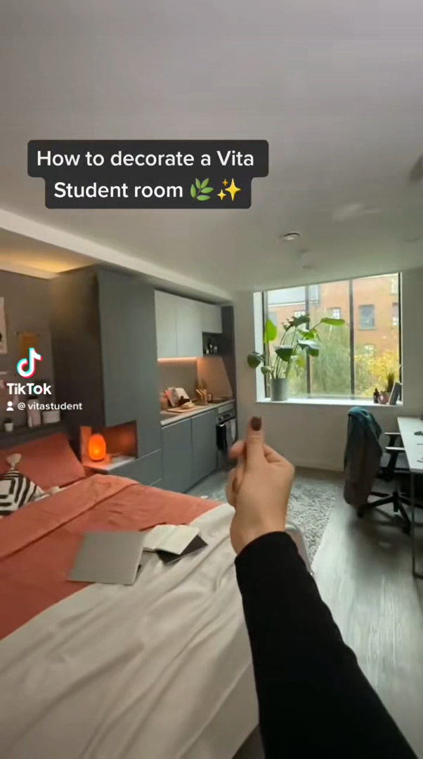 A cozy student room with a bed, a small kitchen area, and a desk near a large window. The hand in the foreground points at the desk adorned with books on education, a plant, and a laptop. Text overlay reads, "How to decorate your Vita Student room while studying.