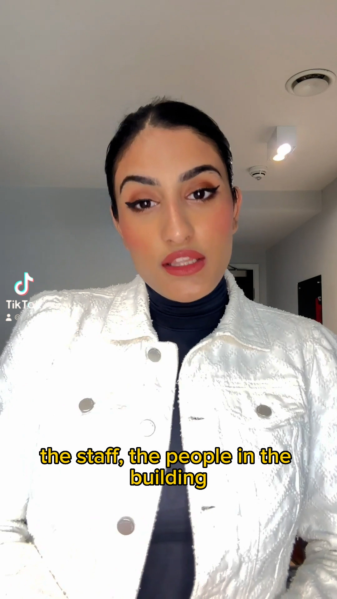 A person with dark hair tied back wears a white jacket and dark top, appearing to speak while standing indoors. Yellow subtitles read: "the staff, the people in the building." A TikTok logo is visible on the left, as if sharing insights about studying abroad in India.